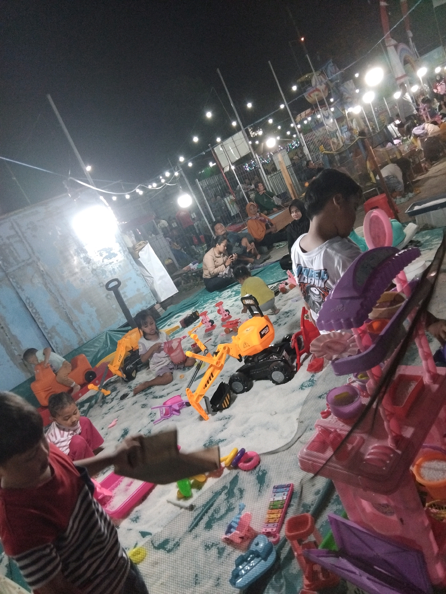 The image depicts a lively outdoor scene at night, likely a community event or playground in Sedati Agung, Indonesia. String lights illuminate the area, casting a warm glow on numerous children engaged in play. In the foreground, several children are seated on a large, light-colored mat spread over the ground, surrounded by a variety of toys. Prominently featured are construction toys, including a bright orange toy excavator and a black toy truck. Other toys visible include colorful plastic kitchen playsets, a toy xylophone, and various small plastic accessories like cups and bowls.
In the midground, more children are playing. One young girl in a pink and white striped shirt is actively playing with a pink basket. Another child, partially obscured, is sitting behind an orange toy vehicle. Further back, a group of adults and children are seated, some watching the children play and others seemingly interacting amongst themselves. The background reveals a bustling atmosphere with more lights and structures, possibly indicating a fairground or carnival setting. The overall impression is one of joyful, communal activity under the night sky in an Indonesian community space.