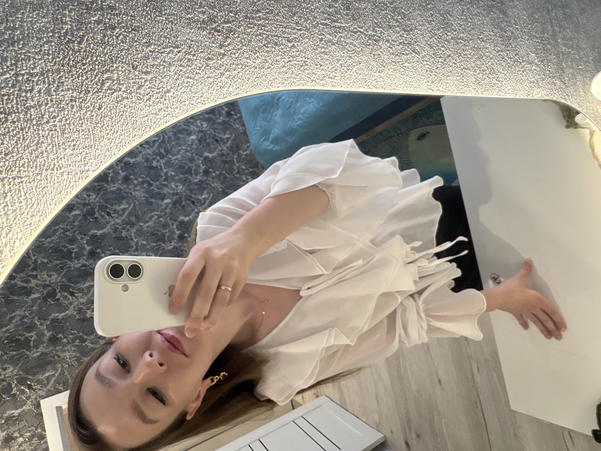 A person with long brown hair is taking a selfie in a mirror. The person is wearing a white ruffled shirt and jewelry. The mirror has a light border and reflects a portion of a room with a blue cushion and gray marble-patterned surface. The mirror's edges are irregular, and the reflection includes a white object. The location is not specified within the visual information.