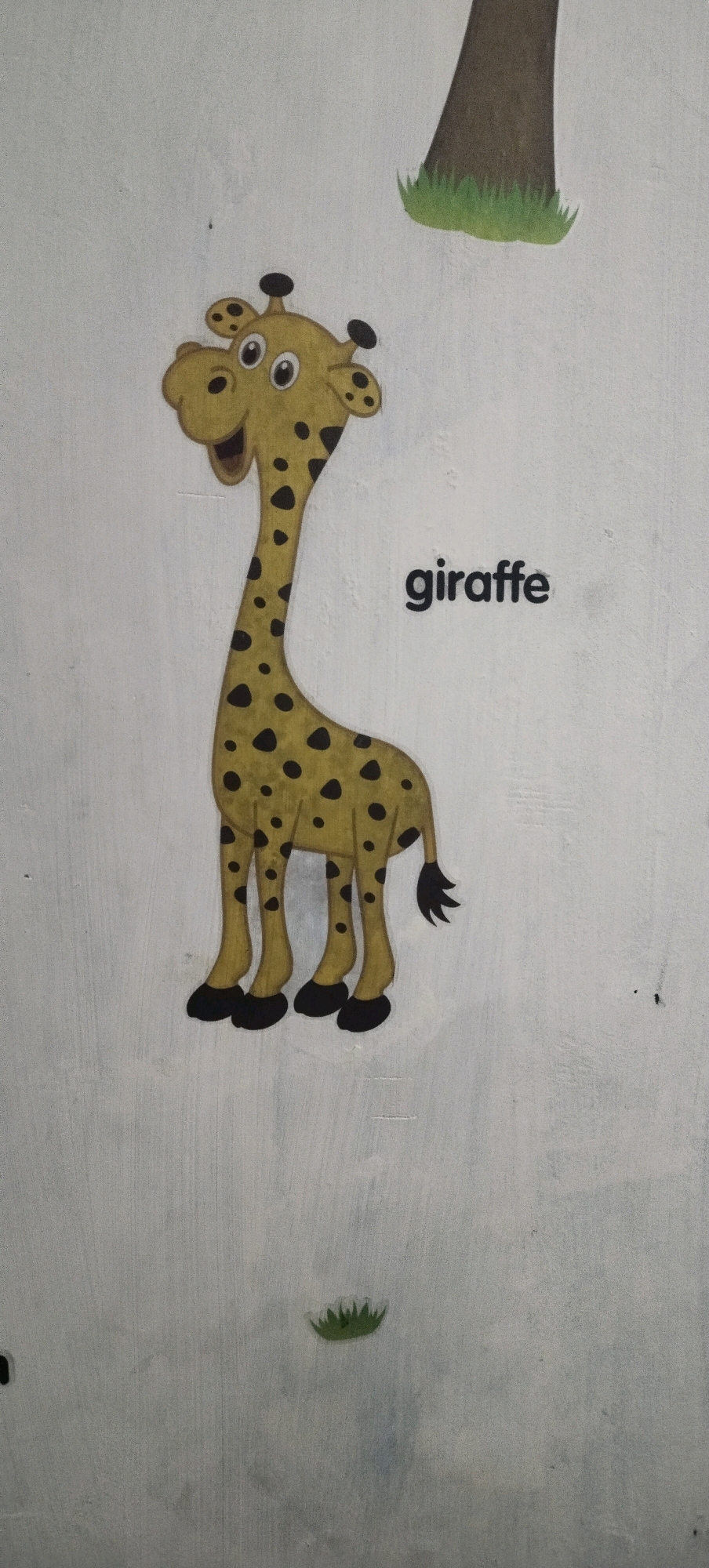 An illustration of a cartoon giraffe is affixed to a light-colored wall in what appears to be a children's room or classroom in Daleman, Indonesia. The giraffe is depicted in a standing position, facing left, with a spotted yellow body, black spots, and black hooves. Its head is tilted slightly, and its mouth is open in a smile. Above and to the right of the giraffe, the word "giraffe" is printed in black, sans-serif font. To the upper right of the giraffe, the base of a tree trunk with green grass at its bottom is visible, also affixed to the wall. The wall itself has a textured surface with visible vertical lines. In the lower center of the image, a small clump of green grass is also affixed to the wall. The lighting suggests an indoor environment, possibly with ambient light from a window or overhead fixture.