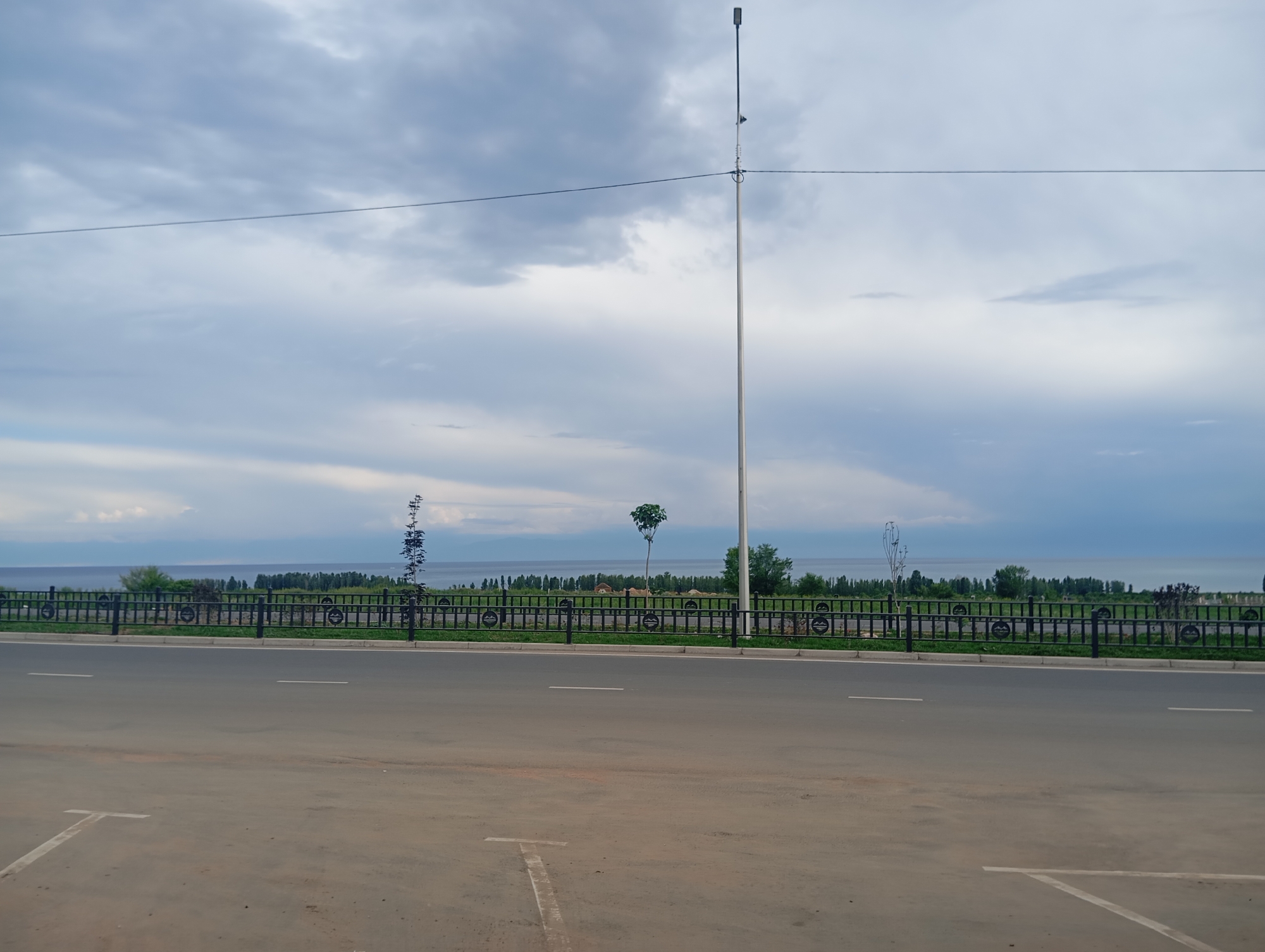 The image depicts a serene outdoor scene. In the foreground, a paved road with painted white lines suggesting parking spaces is visible. A black metal fence runs parallel to the road, separating it from a grassy area. Beyond the fence, a body of water, likely a lake, stretches across the horizon. The far shore is lined with a dense array of trees.
The sky is predominantly overcast with dramatic, textured clouds in shades of grey and white, suggesting either early morning or late afternoon light. A tall, thin lamppost stands prominently on the right side of the frame, its light fixture positioned at the very top. A thin wire stretches horizontally across the sky, connecting to the lamppost.
There are several trees visible on the grassy area between the fence and the water, including a distinctive, small tree with a rounded canopy located near the center. The overall atmosphere is peaceful and tranquil. The weather appears to be mild, with no obvious signs of rain or strong winds. The location cues, such as the trees and the body of water, are consistent with a lakeside setting in Kyrgyzstan. No people are visible in the image.
