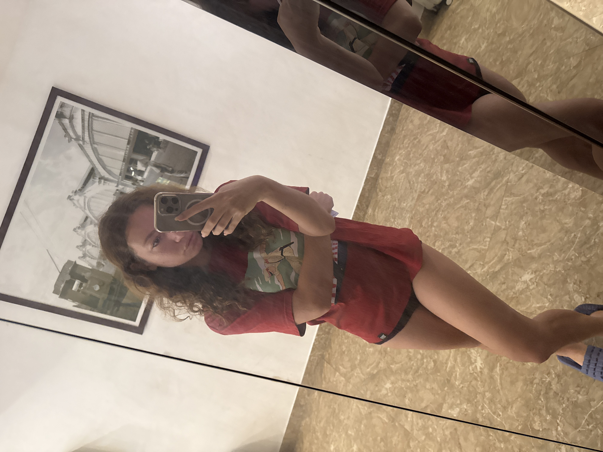 A young woman with curly brown hair is taking a mirror selfie in what appears to be a hotel room. She is wearing a red t-shirt and black underwear. Her arms are crossed, and she is holding her phone in her right hand. In the background, there is a framed picture on the wall, and a full-length mirror. The floor is tiled with a marble pattern. The room appears to be well-lit. There is no indication of the location being in Hanoi, Vietnam.