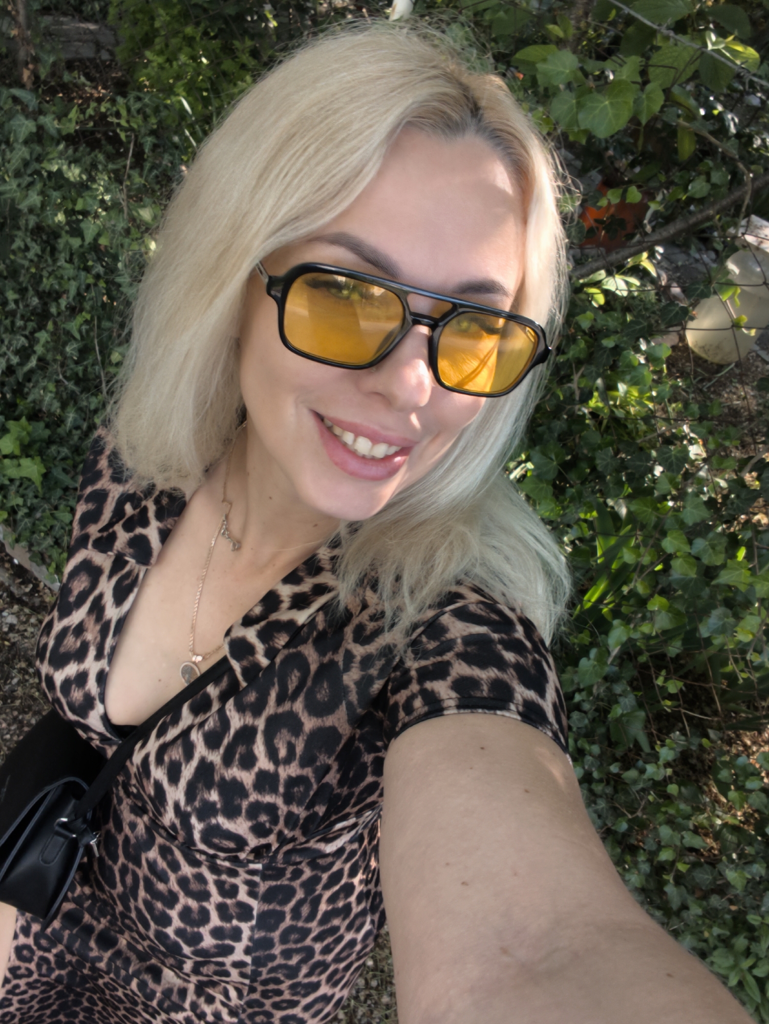 A blonde-haired woman wearing yellow-tinted glasses and a leopard-print dress takes a selfie. A black purse is slung over her shoulder. The background features greenery, suggesting an outdoor location. The precise location within Sevastopol, Ukraine, cannot be determined from this image.