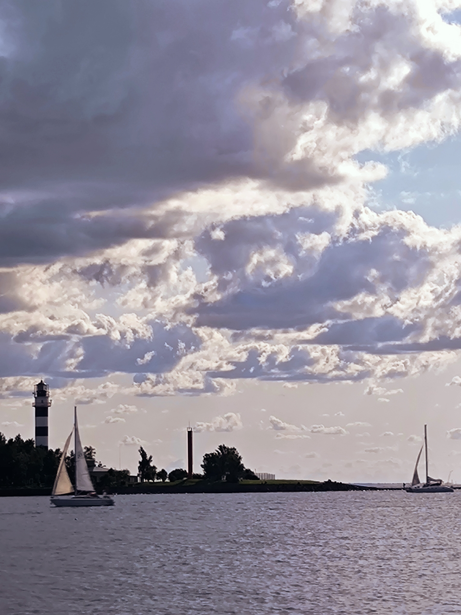The image depicts a scene on the Daugava River in Riga, Latvia, with the distinctive lighthouse of Daugavgriva in the background. The sky is filled with dramatic, textured clouds, ranging from dark, brooding purples to lighter, illuminated whites. Two sailboats are visible on the water. The larger sailboat in the foreground is white with a beige sail, positioned towards the left side of the frame. Further in the distance, to the right, a second, smaller sailboat with a tan sail is partially visible. On the shoreline, the Daugavgriva lighthouse, a tall, striped structure (black and white), stands prominently. To its right, a smaller, red lighthouse is also visible. The water is a deep, slightly rippled blue-grey, reflecting the muted tones of the sky. The overall impression is of a somewhat dramatic, overcast day over a maritime landscape.