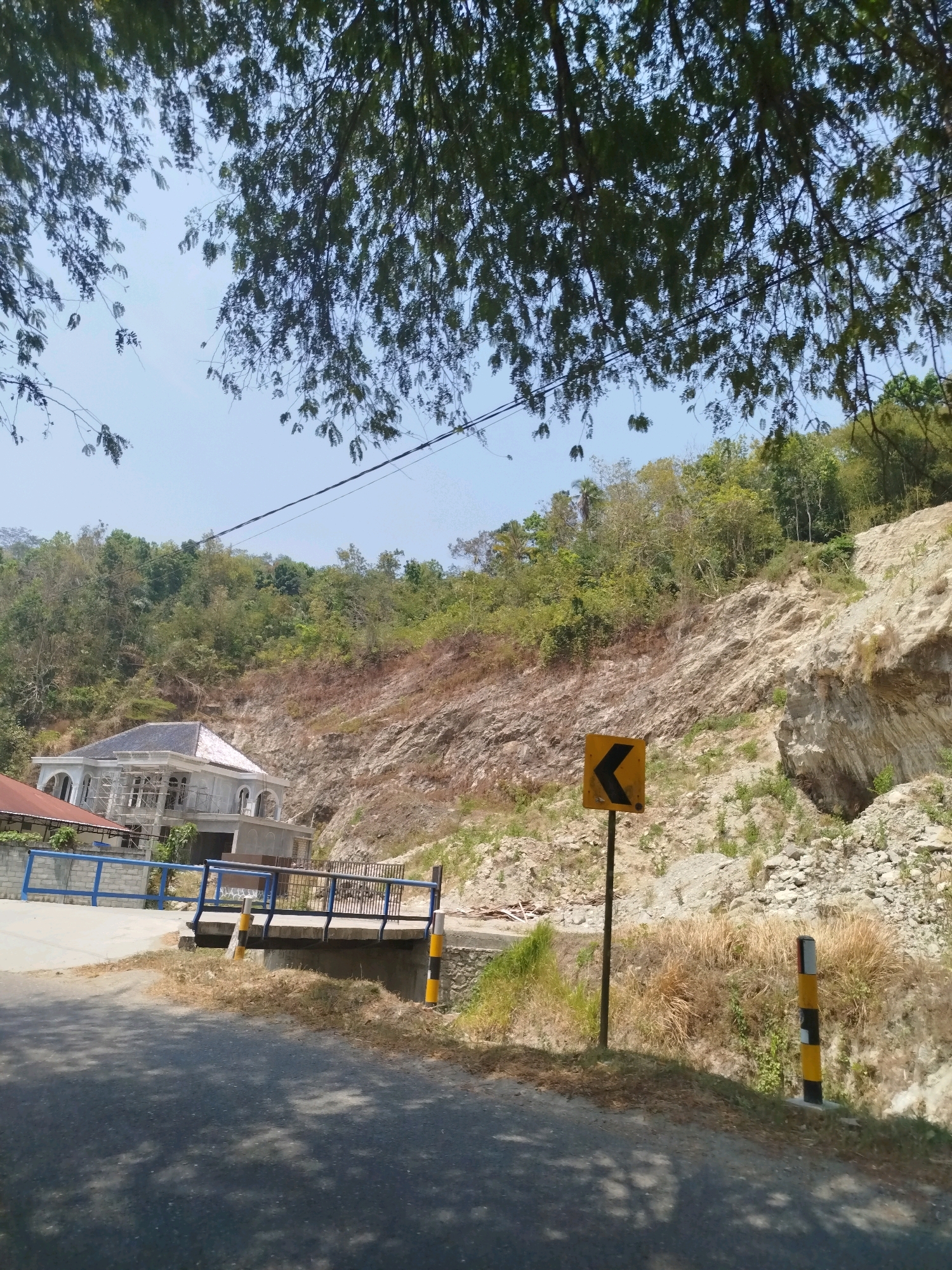 In Kutosari, Indonesia, a road winds through a mountainous terrain. The scene is dominated by a steep, rocky hillside covered in sparse vegetation. A partly constructed building, appearing to be a multi-story house with arched windows and a visible scaffolding structure, is nestled into the hillside, partially obscured by the surrounding trees. The building has a dark-tiled roof and a light-colored facade.
In the foreground, the road surface is visible, with shadows from overhead trees casting dappled patterns. A bridge with a blue railing crosses a small ravine or ditch, with yellow and black striped posts on either side. Adjacent to the road, a yellow directional sign with a black arrow pointing left is mounted on a pole. Another black and yellow striped post stands further along the roadside. The sky is clear and blue, with the upper branches of leafy trees framing the top of the image. The overall impression is one of a rural or semi-rural environment undergoing development, with the natural landscape playing a significant role. There are no people visible in the image.