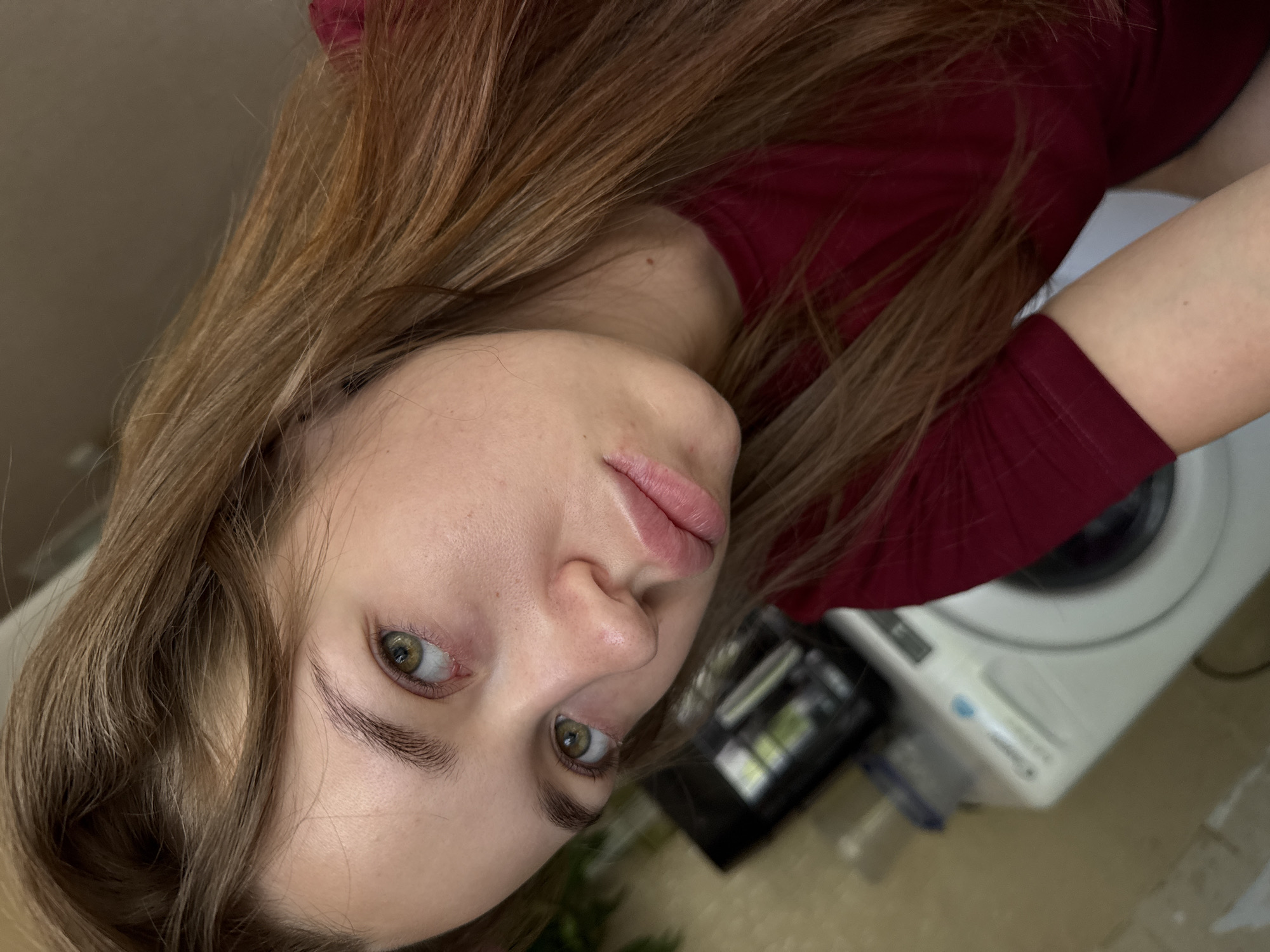 A medium shot selfie shows a young woman with long, auburn hair, her head tilted to the side. She has fair skin, light brown eyes, and full lips. Her expression is neutral, and she is wearing a dark red t-shirt. In the background, a white washing machine is partially visible, suggesting an indoor setting, possibly a laundry room or bathroom. The overall lighting is soft, creating a natural feel to the image. There is no visible indication of the location being specifically in the city of Chaikovsky, Russia, as there are no identifying landmarks or context provided in the image.