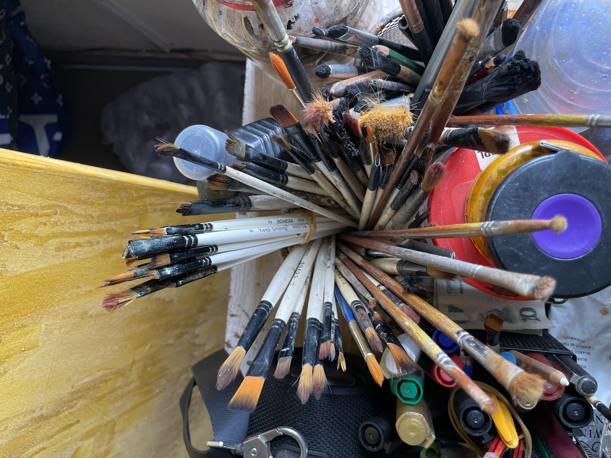 The image is a close-up, high-angle shot of an assortment of art brushes, many of which appear well-used, crammed into a container. The background consists of a textured, yellow surface, possibly a canvas or wall, with a blurred dark area to the right. The brushes have varying bristle types and colors, including white, black, and orange, and many have dried paint residue on them. Some of the handles are white with black tips, while others are made of wood. Other art supplies, such as what appear to be bottle caps of paint and possibly a roll of tape, are visible interspersed with the brushes. The overall impression is one of a working artist's studio or supply area. The location in Ado, Nigeria, is not visually indicated in the image.