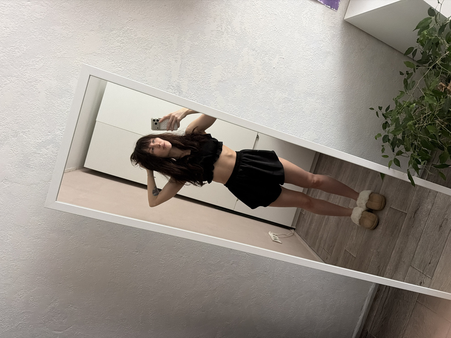 The media file displays a full-length mirror selfie of a woman, likely taken indoors in Perm, Russia. The woman, with long dark hair and bangs, is seen reflected in a large, white-framed mirror that is tilted at an angle. She is wearing a black ruffled crop top, black shorts, and tan fuzzy slippers. Her left arm is bent, with her hand near the back of her head, while her right hand holds a smartphone, presumably taking the picture. Her expression is neutral as she looks towards the phone.
The background reflected in the mirror includes light-colored walls, minimalist white furniture (possibly a dresser or cabinet), and a light wood or laminate floor. To the right of the mirror, an indoor plant with green foliage is visible, partially obscured. The walls outside the mirror's frame exhibit a textured, light grey finish. The lighting suggests it is daytime, illuminating the room evenly. There is no visible text in the image. The overall scene conveys a casual, domestic moment.
