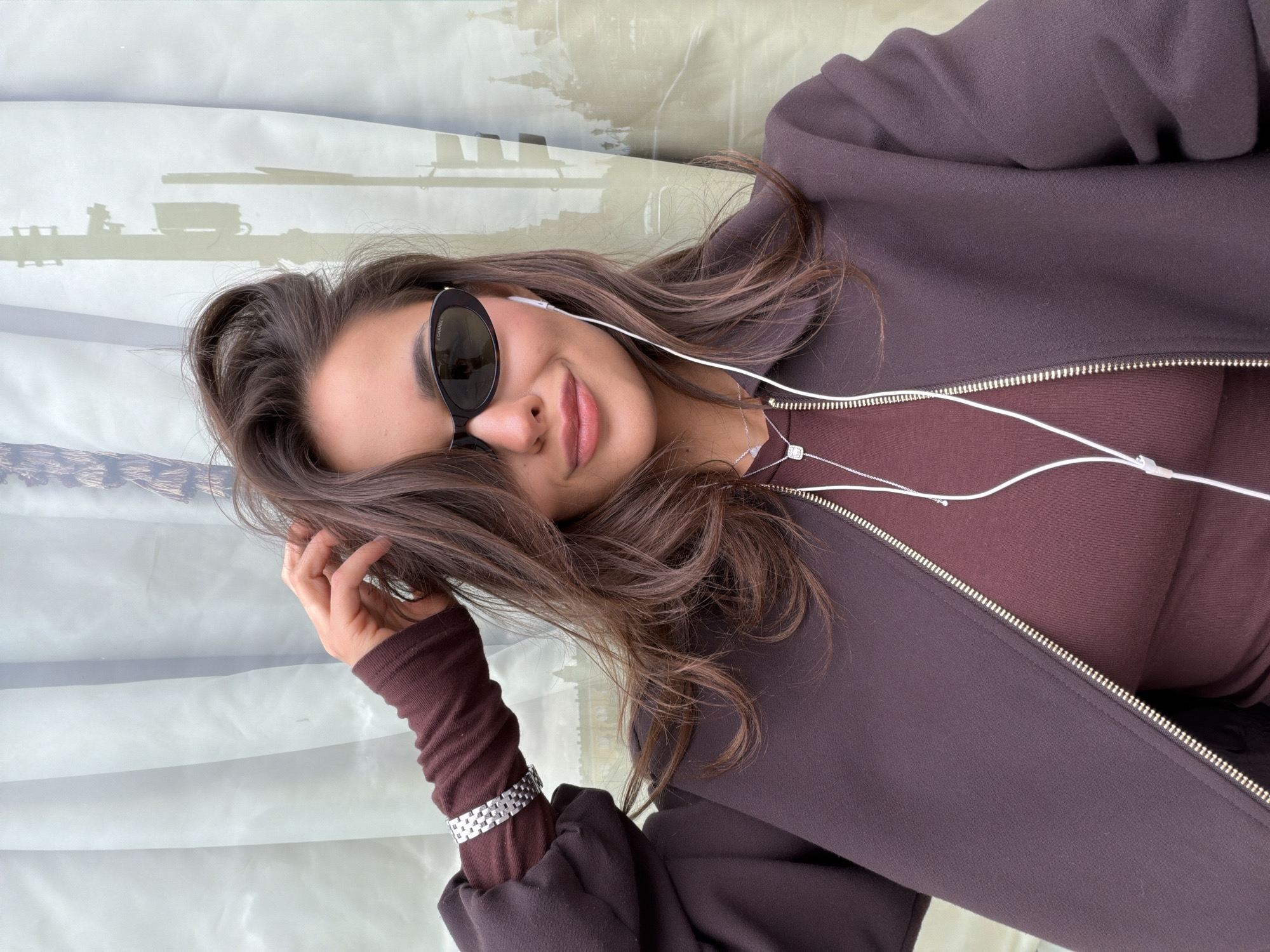 A young woman is depicted in a medium close-up, portrait-oriented photograph, angled diagonally. She has long, wavy brown hair, dark eyebrows, and a relaxed expression with a soft smile on her lips. She wears dark, cat-eye sunglasses, with the brand name "CHANEL" visible on the right arm. White wired earbuds are inserted into her ears, with the wire extending down over her clothing.
She is dressed in a dark brown, possibly hooded, zippered jacket or hoodie, worn over a lighter, ribbed brown top. A delicate silver necklace with a small, rectangular, diamond-studded pendant adorns her neck. On her left wrist, a silver link bracelet is visible, worn over a close-fitting, dark brown sleeve. Her right hand is gently placed near her hair.
The background is softly blurred, showing abstract light-colored shapes and hints of an outdoor urban environment, likely viewed through a window. Closer examination of the provided crops reveals discernible architectural silhouettes in the background that strongly suggest the distinctive multi-domed spires of St. Basil's Cathedral, consistent with the provided location context of Moscow, Russia. The scene is brightly lit, indicating daytime.