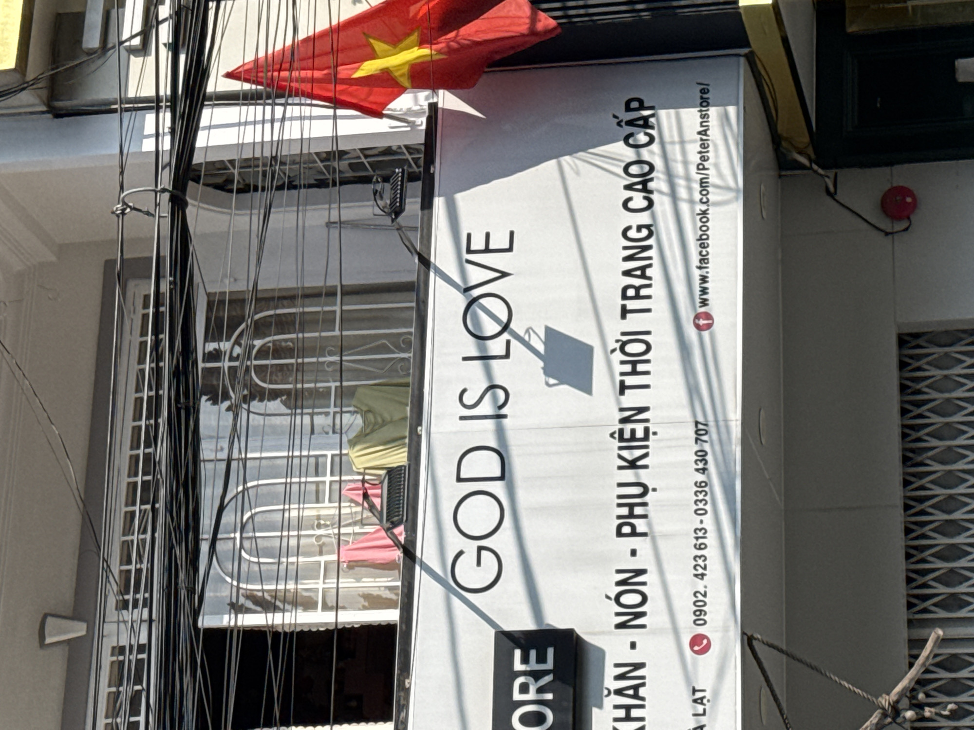 The image displays a bustling street scene in Da Lat, Vietnam, characterized by a detailed view of a building's facade and a prominent commercial sign. A large, white vertical banner dominates the center, featuring bold black text. The English phrase "GOD IS LOVE" is prominently displayed. Below it, in Vietnamese, is "KHĂN - NÓN - PHỤ KIỆN THỜI TRANG CAO CẤP," which translates to "Scarves - Hats - High-Class Fashion Accessories." The sign also provides contact information: phone numbers "0902. 423 613" and "0336 430 707," along with a Facebook page URL "www.facebook.com/PeterAnstore/," and the partial word "LAT" (likely referring to Da Lat).
Above and to the left of the sign, a red Vietnamese flag with a yellow star is visible, fluttering. The foreground is filled with a dense tangle of black electrical and communication wires that run vertically, partially obscuring the building behind them. The building itself is light-colored, featuring multiple windows, some with decorative white security grilles. Through one such window or balcony, a yellow and a pink garment can be seen hanging to dry. To the right of the sign, a dark green or black roll-down shutter or door is visible, and a small red circular object, possibly an alarm or sensor, is mounted on the wall. The scene is brightly lit by strong sunlight, casting distinct shadows on the sign and building elements, indicating a clear, sunny day. No people are visible in the frame.