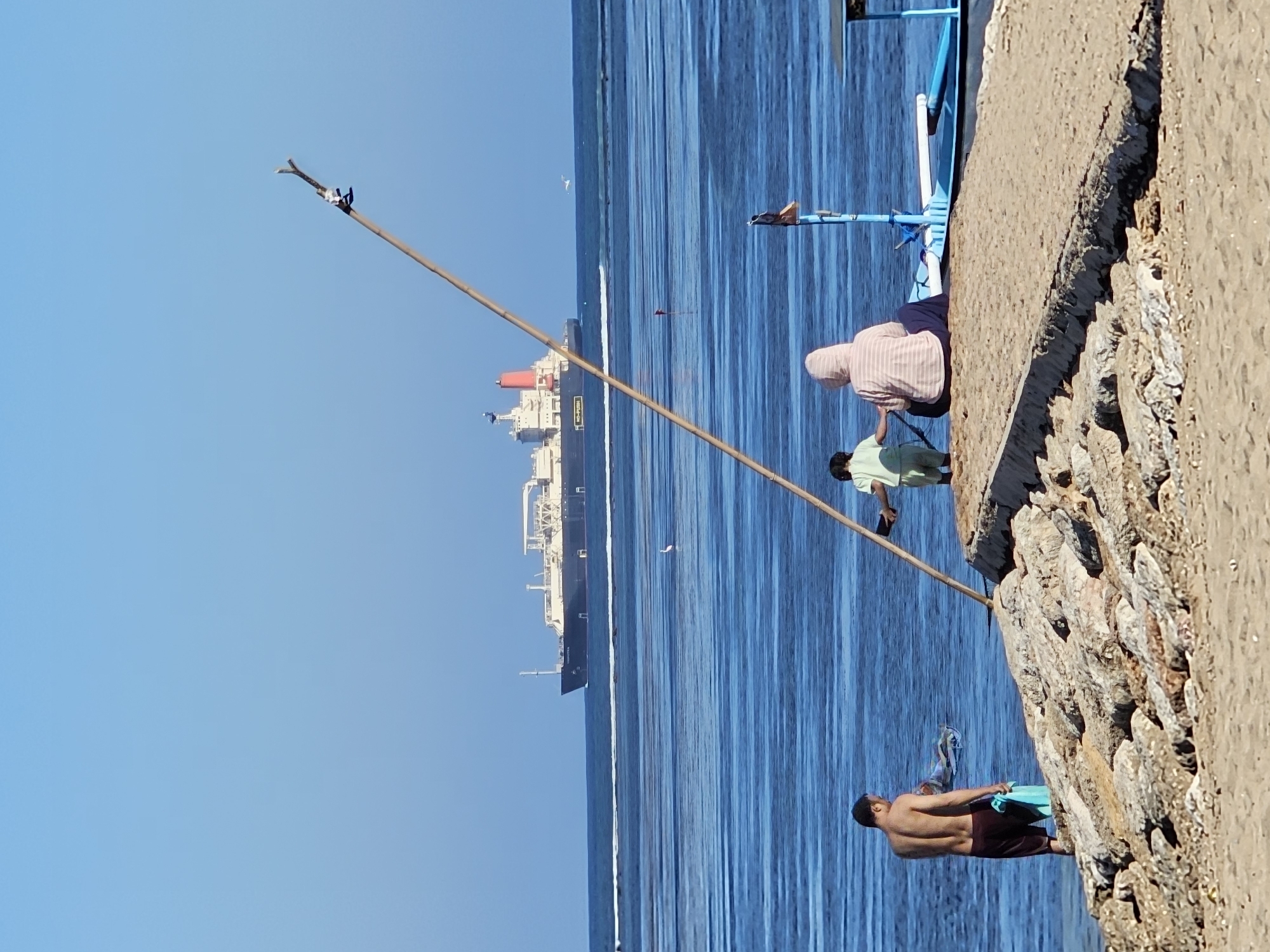 The image depicts a sunny daytime scene at a beach in Denpasar, Indonesia. In the foreground, a man stands partially in the water, holding a light blue towel. To his right, on a concrete jetty, two people are seated, facing away from the camera towards the ocean. One is a child wearing a green shirt and shorts, and the other is an adult wearing a pink and white striped hooded jacket and dark pants. A blue and white boat is docked beside the jetty. A tall, thin wooden pole with a small white object tied to it stands diagonally in the frame, in front of the seated figures. In the middle ground, a large cargo ship is visible on the horizon, sailing across the blue ocean. The water exhibits calm, rippling patterns. The shoreline is composed of sand and rocks. The sky is clear and blue.