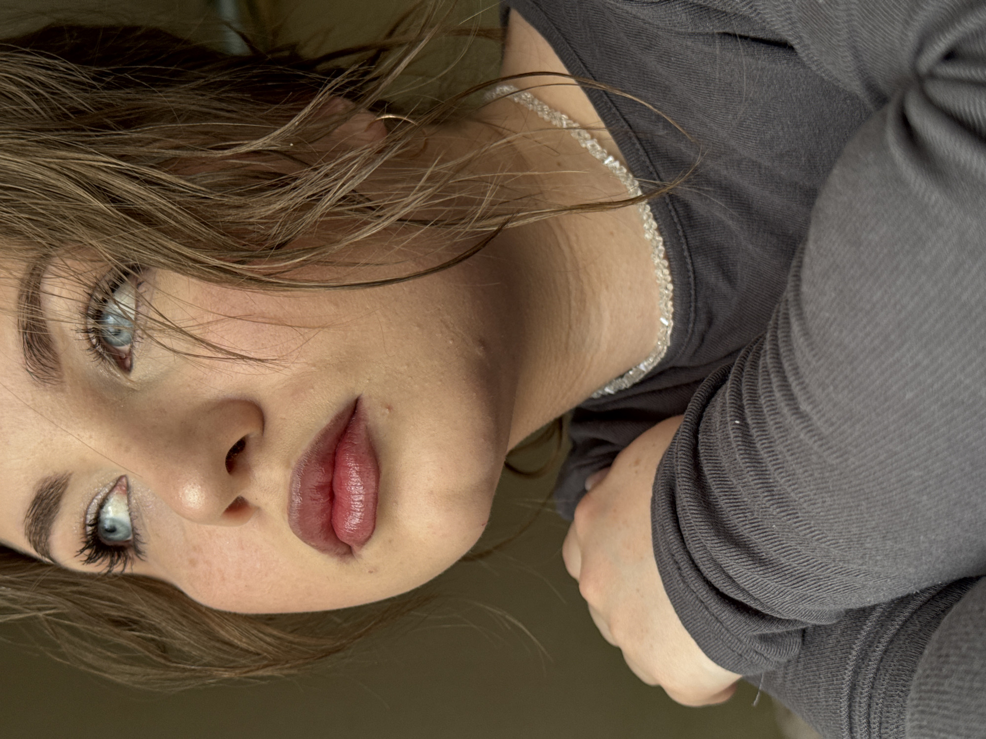 A close-up shot shows a woman's face, angled as though she is reclining or looking downward. She has light brown hair with strands across her face, light skin, and blue eyes. She wears makeup, including eyeshadow and lipstick. A silver beaded necklace adorns her neck. She wears a dark gray long-sleeved shirt. A portion of her hand is visible near her shoulder. The background is a muted, nondescript color. There is no evidence to establish the location in Chaikovsky, Russia.