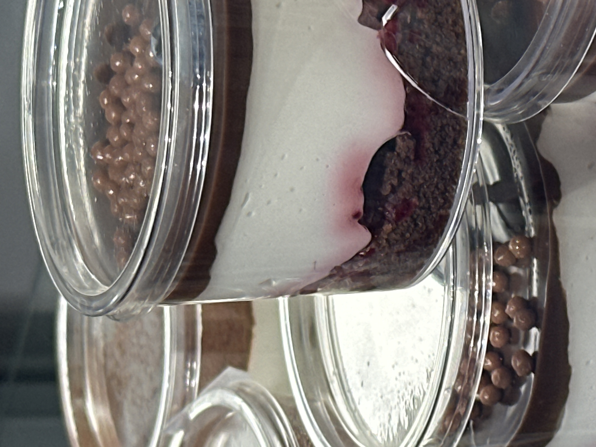 Close-up shot of several clear plastic cups filled with layered desserts, potentially parfaits, stacked. The layers appear to consist of chocolate granules, white cream, and a dark, possibly fruit-based, filling with red streaks. The dessert cups are arranged in a somewhat tilted, overlapping fashion, suggesting a display or storage setup. No people are visible. The specific location within Kazan, Russia, cannot be determined from the image.