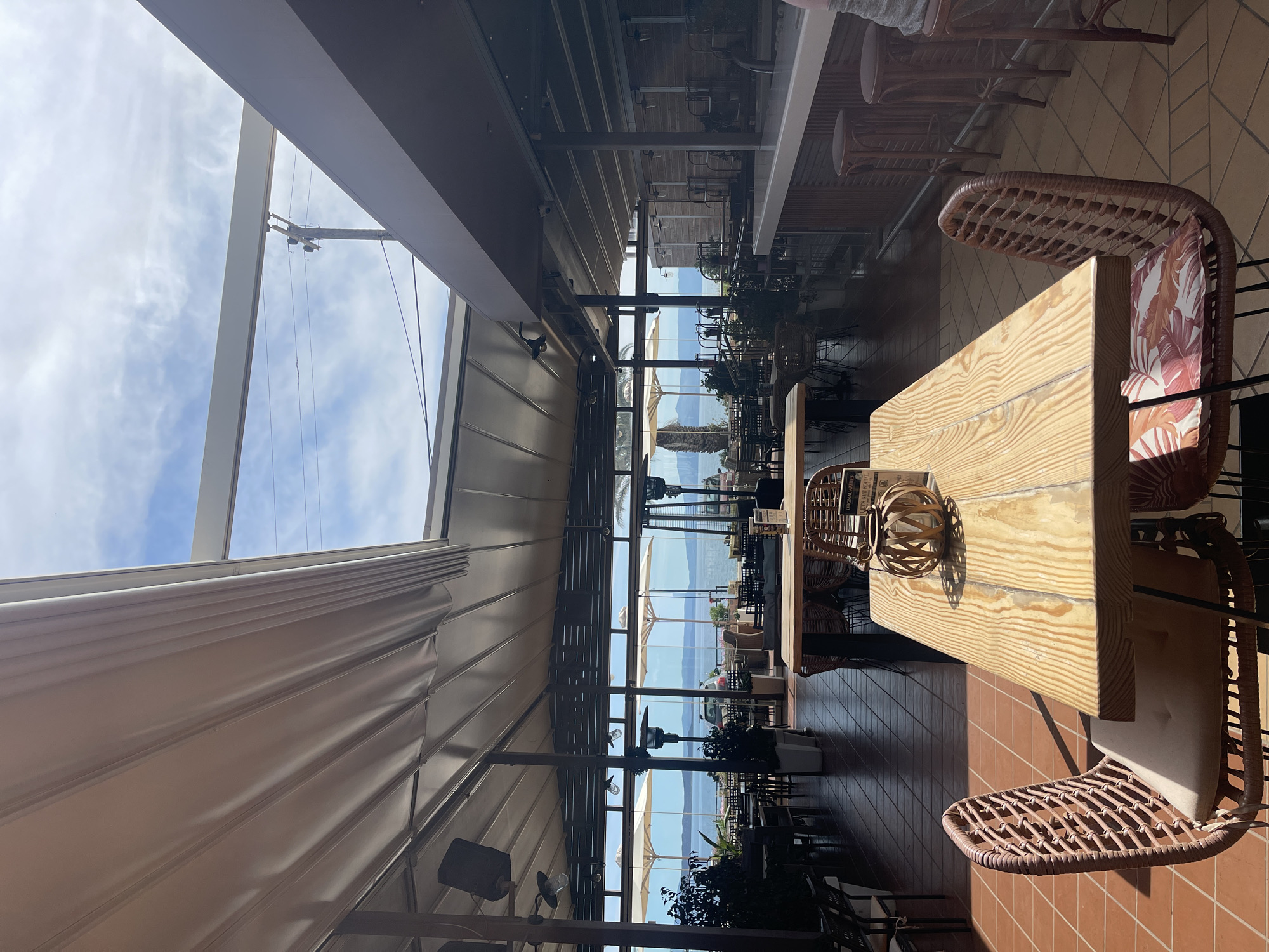 The image depicts the interior of an open-air restaurant or cafe in Nea Makri Municipal Unit, Greece. The space is characterized by a combination of indoor and outdoor elements, with a retractable awning and glass windows offering views of the Aegean Sea. Tables with wicker chairs are arranged on a terracotta-tiled floor, suggesting a casual dining atmosphere. The view extends to the sea, with glimpses of the coastline and the distant island of Euboea. The lighting is bright and sunny, highlighting the wooden tables and wicker furniture. Overall, the scene conveys a relaxed and scenic dining experience in a coastal setting.