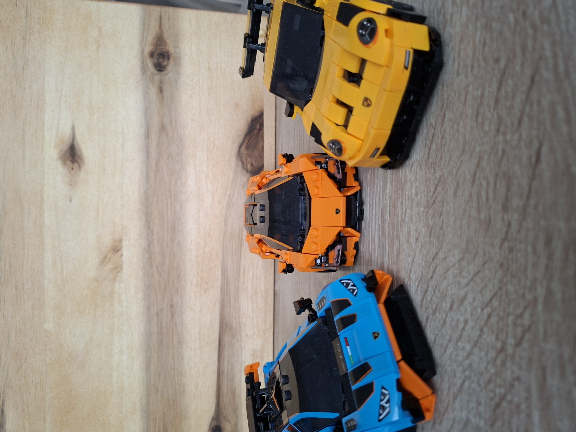 Three toy sports cars are arranged on a wooden surface. From left to right, the cars are blue, orange, and yellow. The blue car is facing forward and slightly to the right, displaying its front grille and headlights. The orange car is positioned in the center, also facing forward. The yellow car is on the right, angled slightly towards the viewer. The background is a textured wooden surface. There is no indication of the city of Baden-Baden, Germany, in the image.