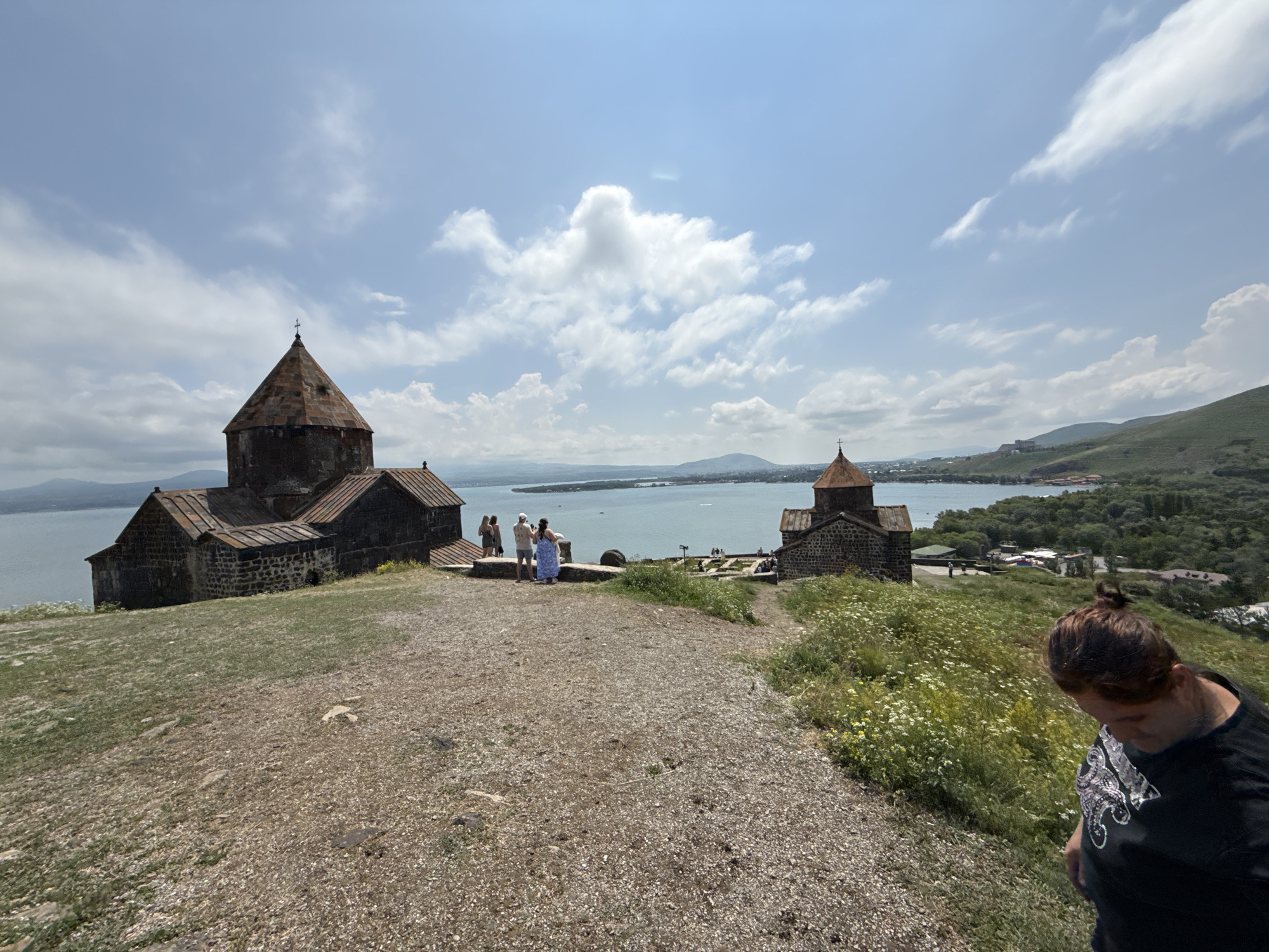 The image captures a view of the Sevanavank monastery complex located on a peninsula in the city of Sevan, Armenia. Two ancient stone churches with conical roofs dominate the left side of the frame, overlooking a vast body of water, likely Lake Sevan. Several individuals are visible standing on the elevated ground near the churches, some appearing to be tourists. The foreground consists of a gravel path and grassy slopes dotted with small white flowers. In the background, a green, hilly landscape recedes towards the horizon, with a few buildings and trees visible. The sky is partly cloudy with scattered white clouds against a blue backdrop. A woman with her hair tied in a bun is partially visible in the lower right corner of the image, looking down.