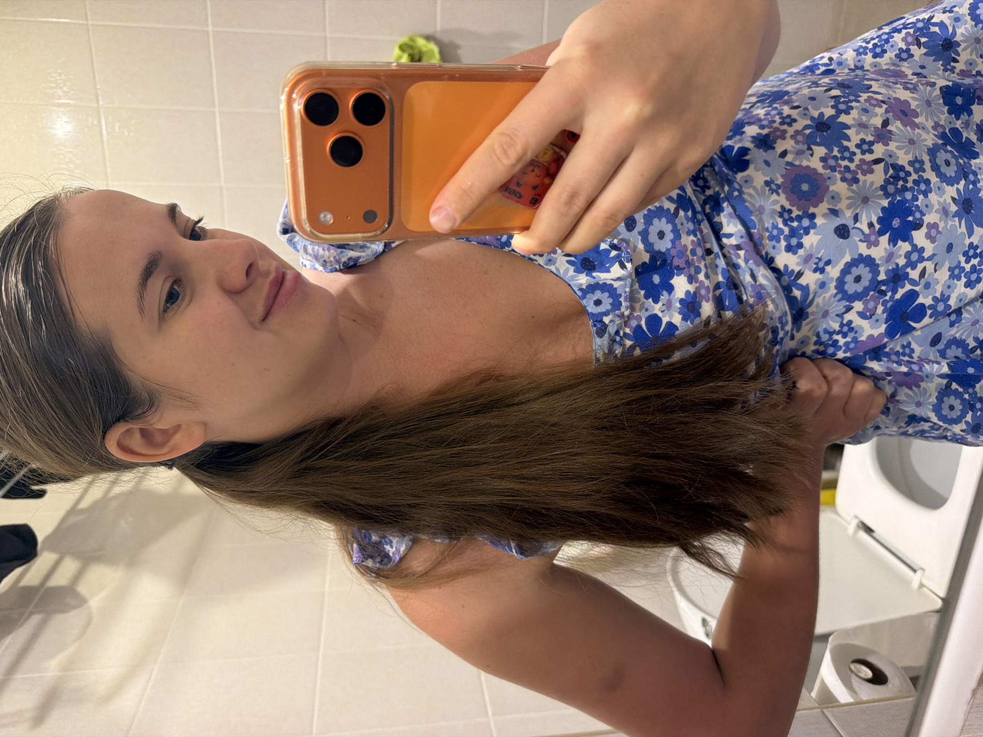 A female subject, appearing to be in her 20s or early 30s, is captured in a mirror selfie within a bathroom setting in the city of Sochi, Russia. Her long, dark brown hair is styled in a low ponytail, with strands near the forehead appearing slightly damp. She is dressed in a sleeveless, V-neck garment, likely a dress, featuring a blue and purple floral pattern on a white base, with ruffled detailing at the shoulders and neckline. The subject holds an orange-cased smartphone, identified as an iPhone by its triple-lens camera system and flash unit, in her right hand, aiming it towards the mirror. Her left hand is placed on her hip, subtly manipulating the fabric of her dress. The bathroom walls are covered with white rectangular tiles. A white toilet with an open lid is visible in the background to the subject's left, alongside a wall-mounted white toilet paper holder. Dark objects are discernible hanging in the upper background, and a small, bright green item is partially visible on the far right wall.