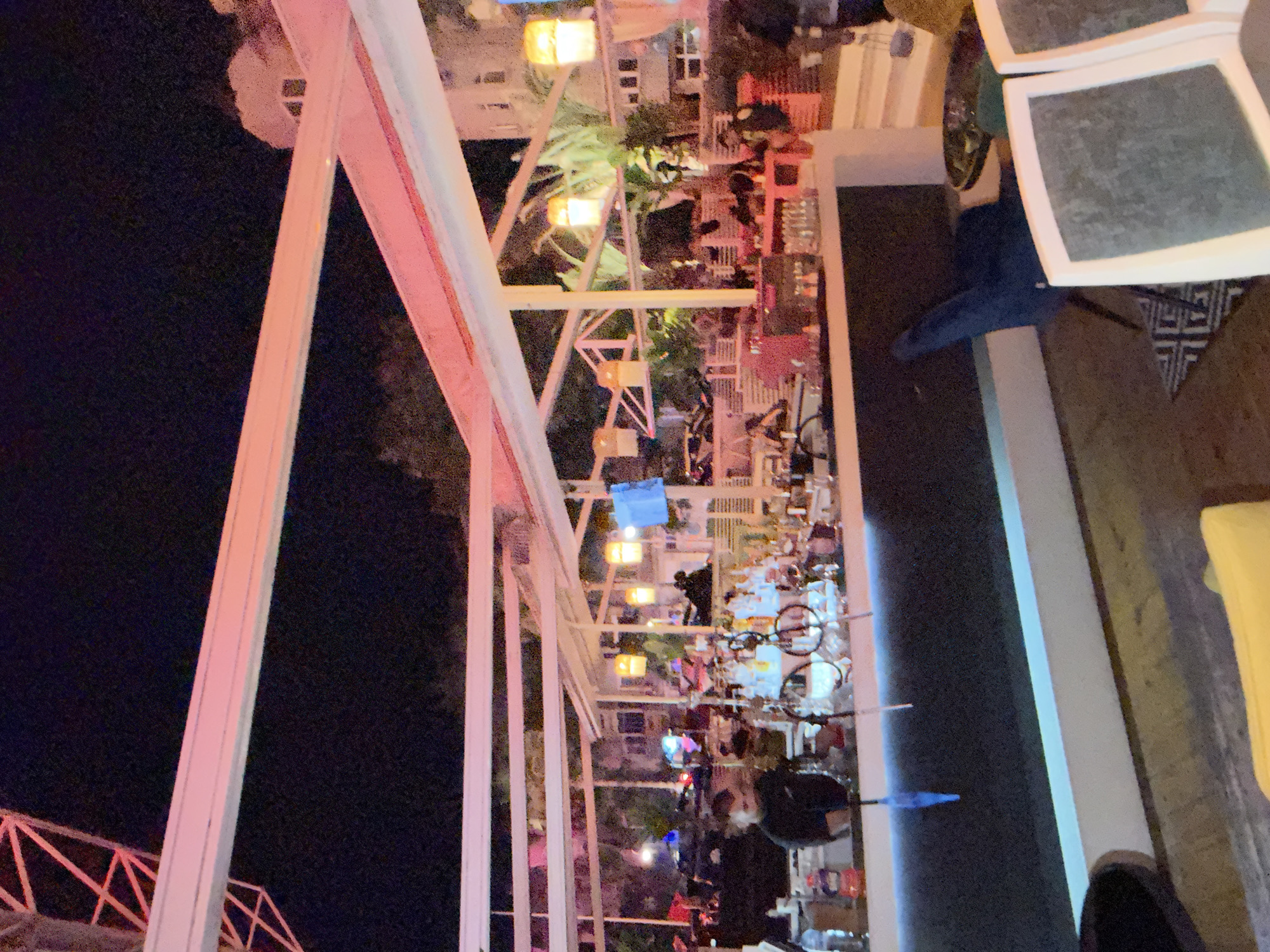 This image depicts an outdoor bar or lounge area at night in the city of Plovdiv, Bulgaria. The scene is dominated by a white pergola or canopy structure illuminated from below with pinkish-red light.
In the foreground, a long, dark-colored bar counter runs horizontally, featuring a horizontal strip of cyan light at its base. Numerous bottles of various alcoholic beverages are arranged on the bar top. A person, identified as a bartender, stands behind the bar, facing right. A blue armchair and a yellow cushion are visible on the left side of the foreground. On the right, parts of chairs with white frames and grey upholstery are seen, along with a small black and white patterned rug under a blue chair.
In the midground, beyond the bar, patrons are scattered throughout the lounge area, some seated at tables with red accents. Several tall potted plants or small trees are interspersed among the seating. Warm yellow-toned hanging lamps are suspended from the pergola structure, providing additional illumination.
The background reveals a dark night sky. On the upper right, a multi-story white building with windows and a distinctive rounded architectural feature is visible. In the upper left corner, a large red metal lattice structure, possibly part of a crane or Ferris wheel, extends upwards against the dark sky. The overall environment suggests an active, illuminated outdoor entertainment venue.