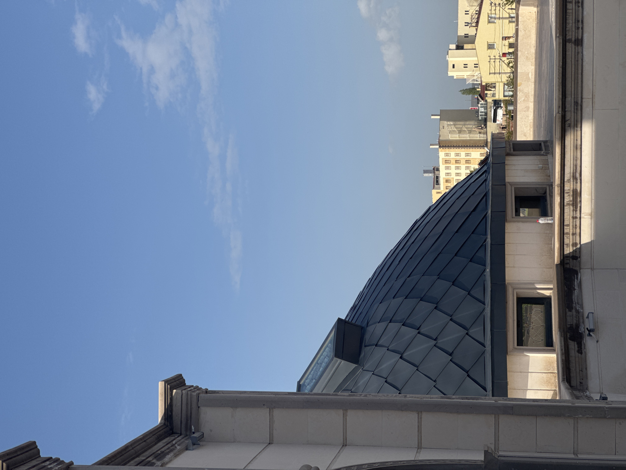 The image shows a low-angle view of building architecture in Ürgüp, Turkey, dominated by a blue-tiled dome and surrounding structures. The left side of the frame is filled with a clear blue sky with scattered clouds. The dome is constructed of individual, grey-blue tiles, forming a scaled pattern. Adjacent to the dome is a building with white stone or stucco facade, small windows, and architectural details. In the background, additional buildings are visible.