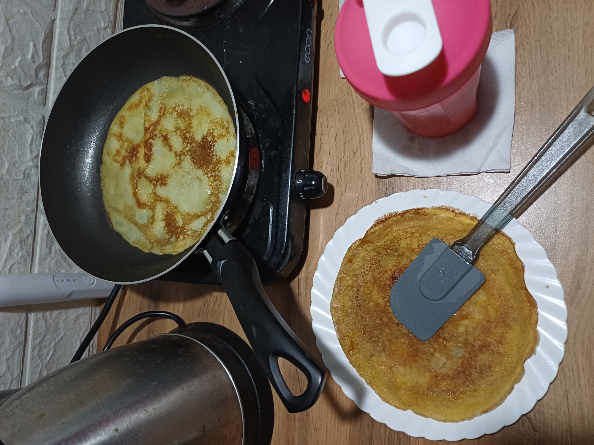 A close-up, overhead shot shows a breakfast scene on a wooden table. On the left, a stainless steel electric kettle stands beside a black electric stovetop. A black frying pan on the stovetop contains a golden-brown crepe, its surface speckled with darker, caramelized spots. To the right of the stovetop, a pink and white water bottle sits atop a white paper napkin. In the foreground, a white plate holds another crepe, with a gray spatula resting on top of it. The background features a white tiled wall. The scene appears to be set in a kitchen or dining area, possibly in a home. There is no indication of the location being in Sochi, Russia.