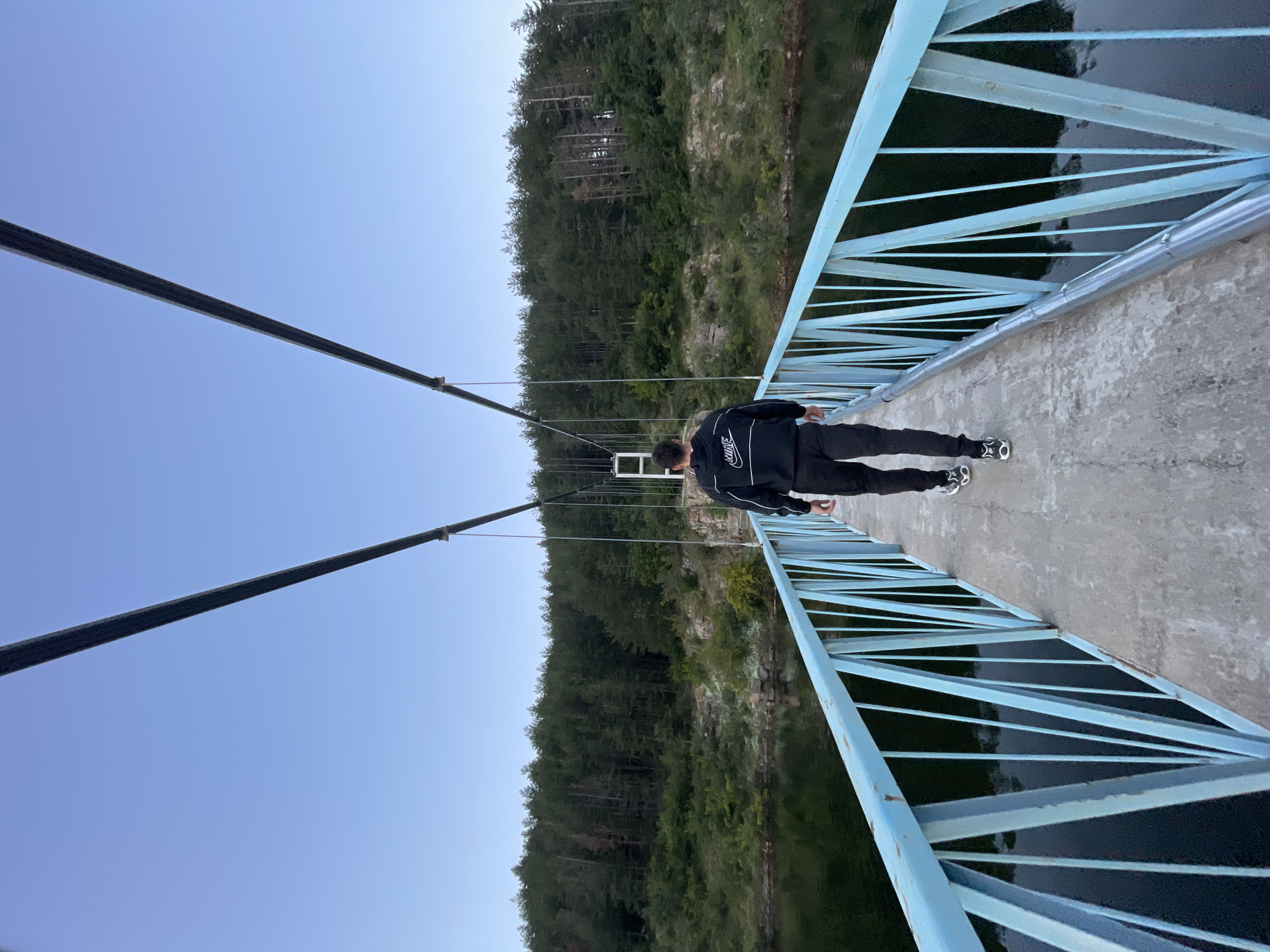 A single male individual, wearing a black Nike hooded sweatshirt with white detailing and black pants, walks away from the camera across a concrete and metal suspension bridge. The bridge features light blue metal railings with a zig-zag pattern of vertical bars. The bridge spans over a dark body of water, likely a river or lake, reflecting the surrounding environment. On the far side of the water, a dense forest of pine trees covers a rocky hillside under a clear, pale blue sky. The location is identified as Suhovo, Bulgaria.