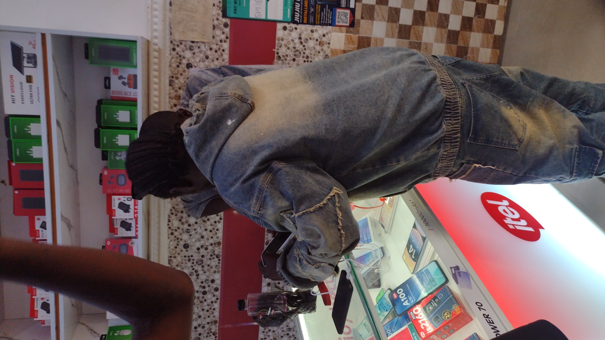 The image shows a person in a denim jacket and pants looking at a display of mobile phones, likely in a retail store. The display case has the "itel" logo prominently displayed, and various itel phones with their boxes are visible. To the left of the display is a shelf unit showcasing boxes of phone accessories and what seems to be a CCTV system box, with brands like "HIT VISION" and "BUDS ACE" visible. The environment includes a wall with a patterned design and a poster with the words "JIUJIO INSTALLMENT". The location is identified from a similar store in the city of Jos, Nigeria. There are no actions or interactions immediately apparent in this image.