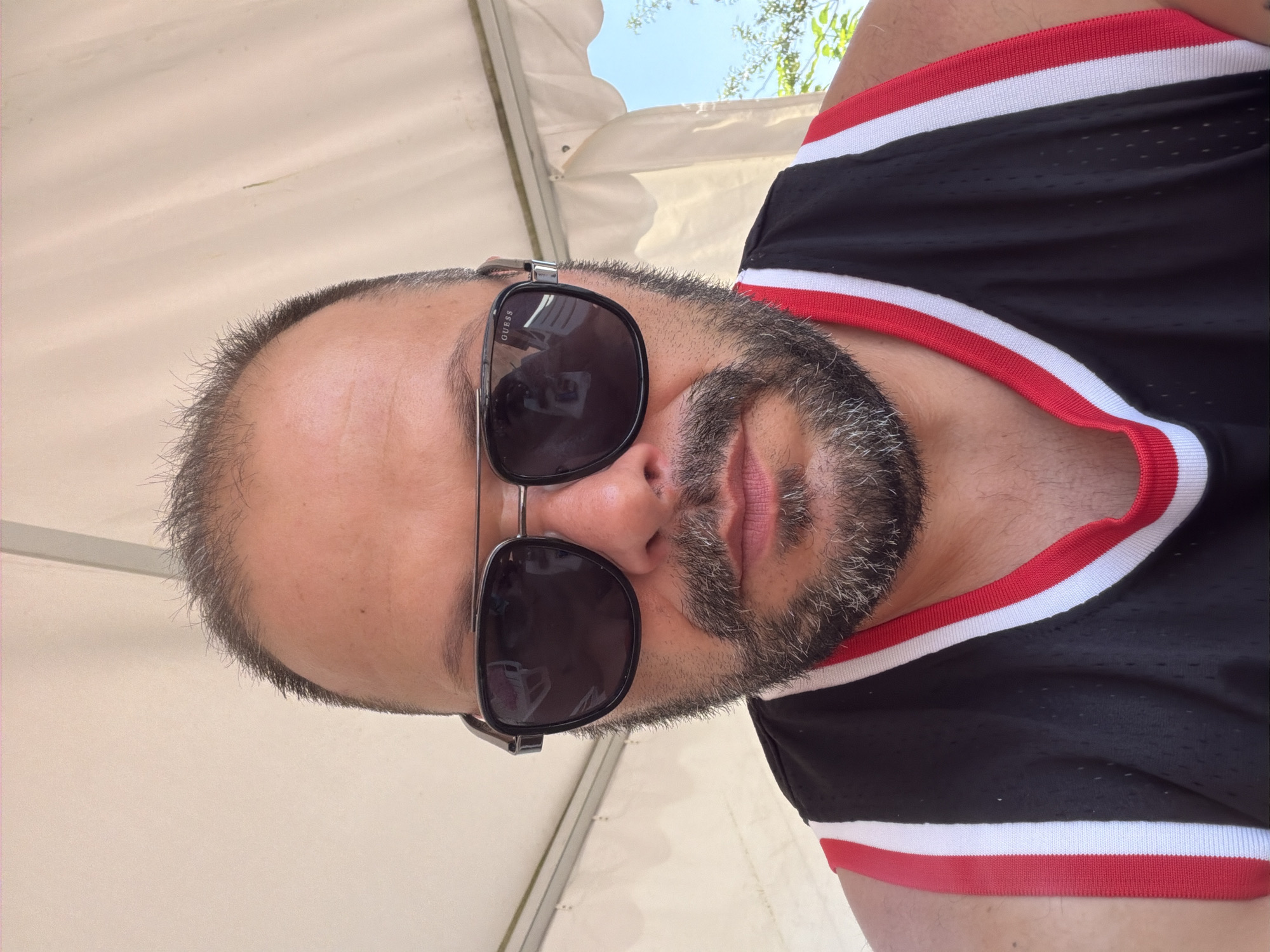 A man is pictured from the chest up, wearing sunglasses and a black tank top with red and white trim. He has a beard and is bald on top with some hair around the sides. The man appears to be outdoors, possibly under a white canopy, with a clear blue sky and some greenery visible in the background. The image was taken in the city of Simeonovo, Bulgaria.