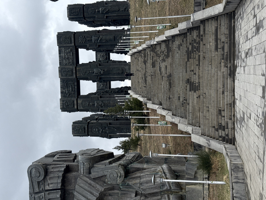 The image depicts the Chronicle of Georgia, a monumental complex situated on a hillside overlooking the Tbilisi Sea in Tbilisi, Georgia. The scene is dominated by a long, wide stone staircase leading upwards towards a massive, dark stone monument under an overcast grey sky.
In the left foreground, a large, dark stone sculpture with intricate, stylized relief carvings and curvilinear forms is prominent, partially cut off by the frame. The central feature is a lengthy, multi-tiered grey stone staircase, appearing wet or stained in patches, ascending towards the monument at the top. This staircase is flanked on both sides by evenly spaced silver-colored lamp posts with inverted saucer-shaped lights, some with green, ornate decorative elements or small bins attached near their bases. Sparse, dry grass and small evergreen trees line the slopes adjacent to the staircase.
At the top of the staircase, the main monument consists of multiple colossal, dark stone or bronze pillars arranged in an open, rectangular formation, forming archway-like structures. These pillars are heavily adorned with detailed bas-relief carvings depicting numerous human figures, historical scenes, and symbolic motifs. One individual, dressed in dark clothing, is visible near the top center of the staircase, walking towards the monument. Another figure is faintly discernible further up and to the right of the monument's entrance. The immediate ground in the foreground is paved with dark grey stone blocks, also exhibiting wet patches.