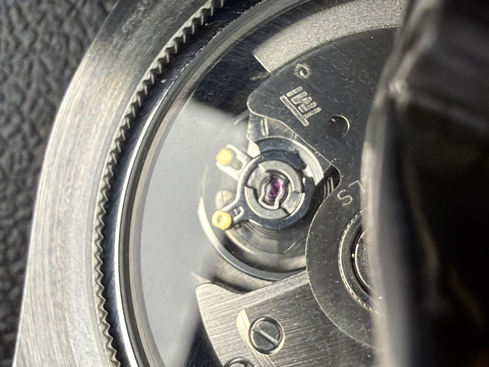 The image shows a close-up view of a watch movement, possibly a Seiko NH35A or similar caliber. The balance wheel, with its characteristic pulse, is visible, along with the pallet fork and escape wheel. Markings on the movement indicate it is a TMI ( a Seiko subsidiary) movement, possibly with "S" marking for the seconds subdial. The watch case appears to be made of stainless steel, with a fluted bezel. The setting is a macro photograph, likely taken in an indoor environment. There are no people or other objects visible in the image. The location in Grapevine, United States of America is not discernible from the image.