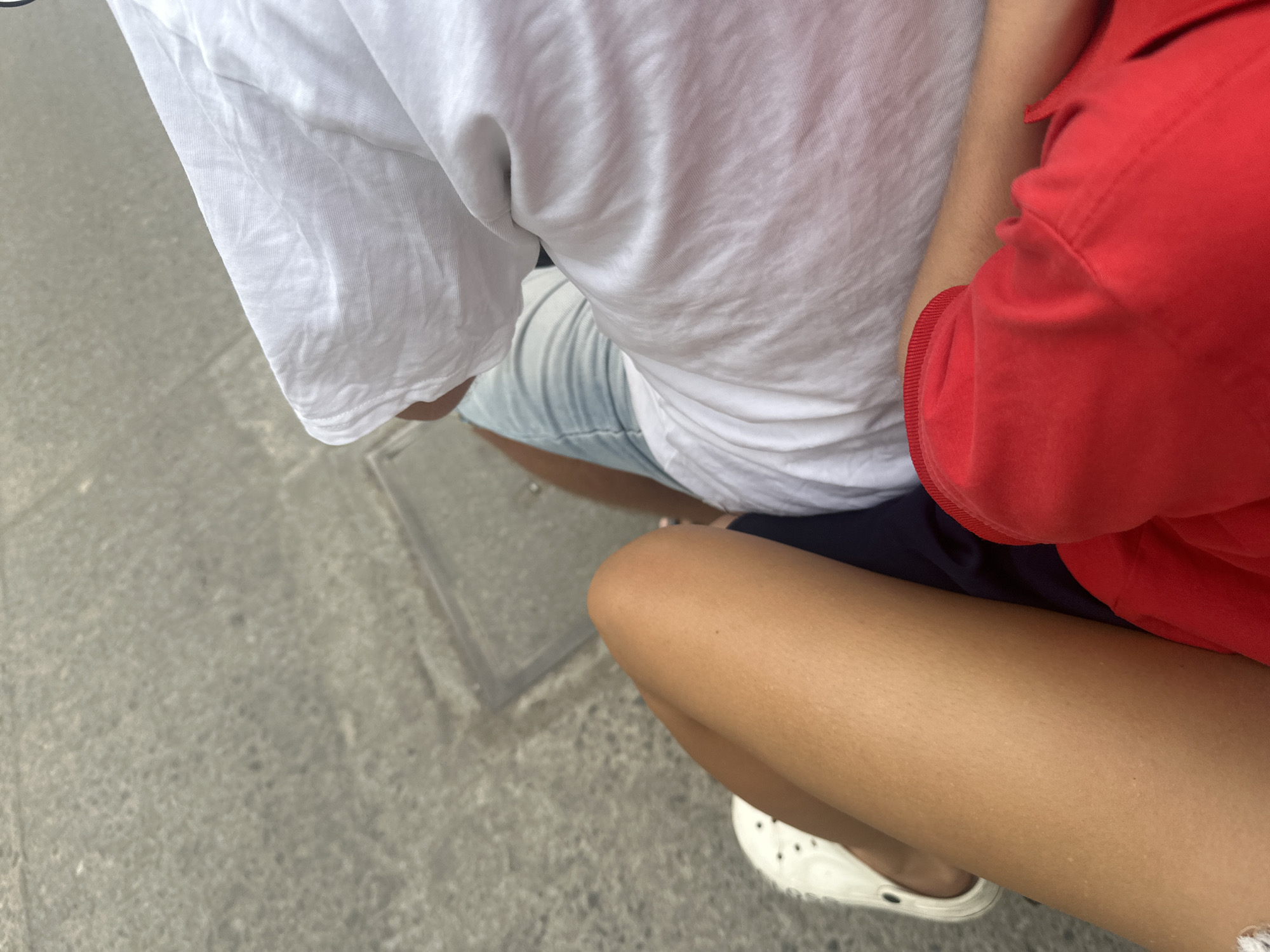 A high-angle shot focuses on two individuals sitting close together outdoors. The person on the left wears a white t-shirt and denim shorts. To their right is another person wearing a red shirt, black shorts, and white Crocs. They are situated on a grey concrete surface. The city of Bac Nha Trang Ward, Vietnam is not discernible in this image.