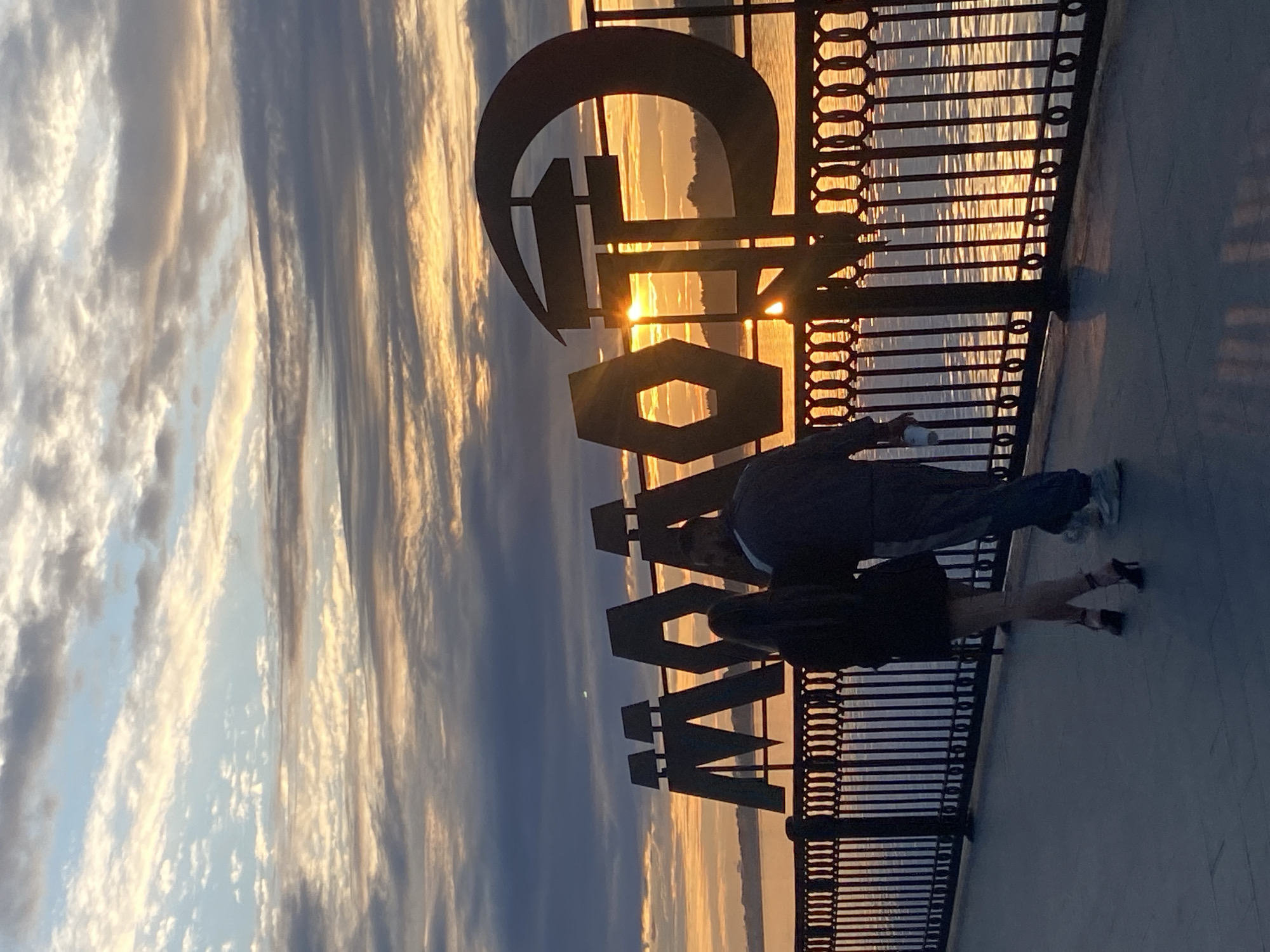 An image captures a couple walking away from the camera along a waterfront promenade in Perm, Russia. The promenade features a black metal fence with decorative circular and vertical elements. Behind the fence, a calm body of water reflects the warm hues of a sunset. Dominating the background is a large, dark metal sculpture spelling out "МООП" in Cyrillic letters. The setting sun casts a golden glow through the letters, creating lens flares. The sky is a canvas of dramatic clouds, with streaks of orange, yellow, and grey against a backdrop of pale blue. The couple, silhouetted against the sunset, are seen from behind. The woman is wearing a black dress and high heels, while the man is in athletic wear, holding a white cup in his right hand. Their figures suggest they are walking together, possibly on a stroll. The overall atmosphere is serene, with the natural beauty of the sunset enhanced by the urban elements of the promenade and the sculpture.