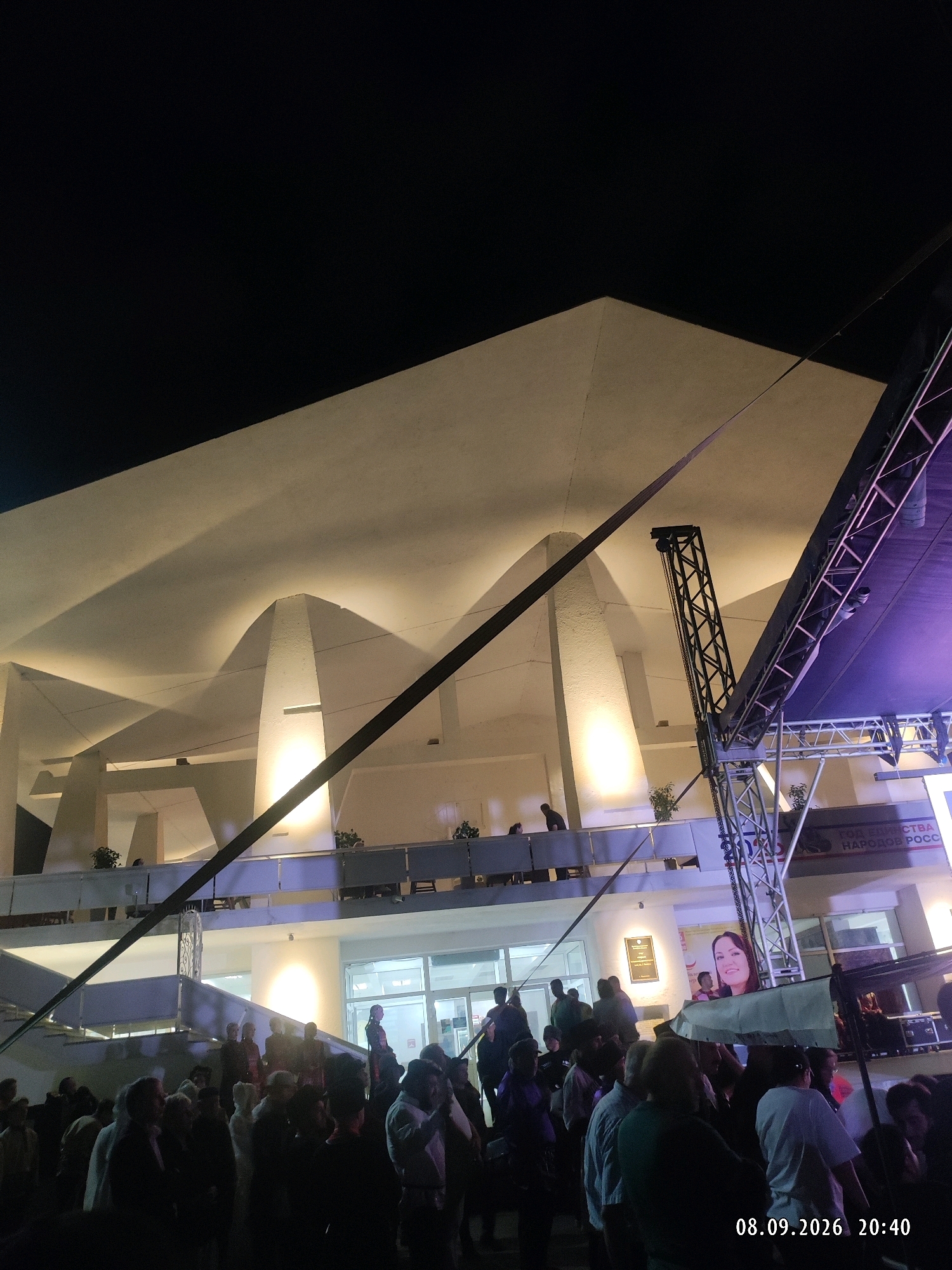 An outdoor nighttime event is captured in this image, likely a concert or public gathering, near the National Congress Palace in Makhachkala, Russia. The building's distinctive, modern architecture features a broad, undulating roofline illuminated by warm spotlights, casting dramatic shadows. In the foreground, a large crowd of people stands, silhouetted against the illuminated building and stage lighting. A metal truss structure, characteristic of stage setups, is prominently visible on the right, with purple lighting emanating from above. A large banner with a woman's portrait is displayed on the building's facade. The timestamp "08.09.2026 20:40" is embedded in the lower right corner of the image, indicating the date and time.