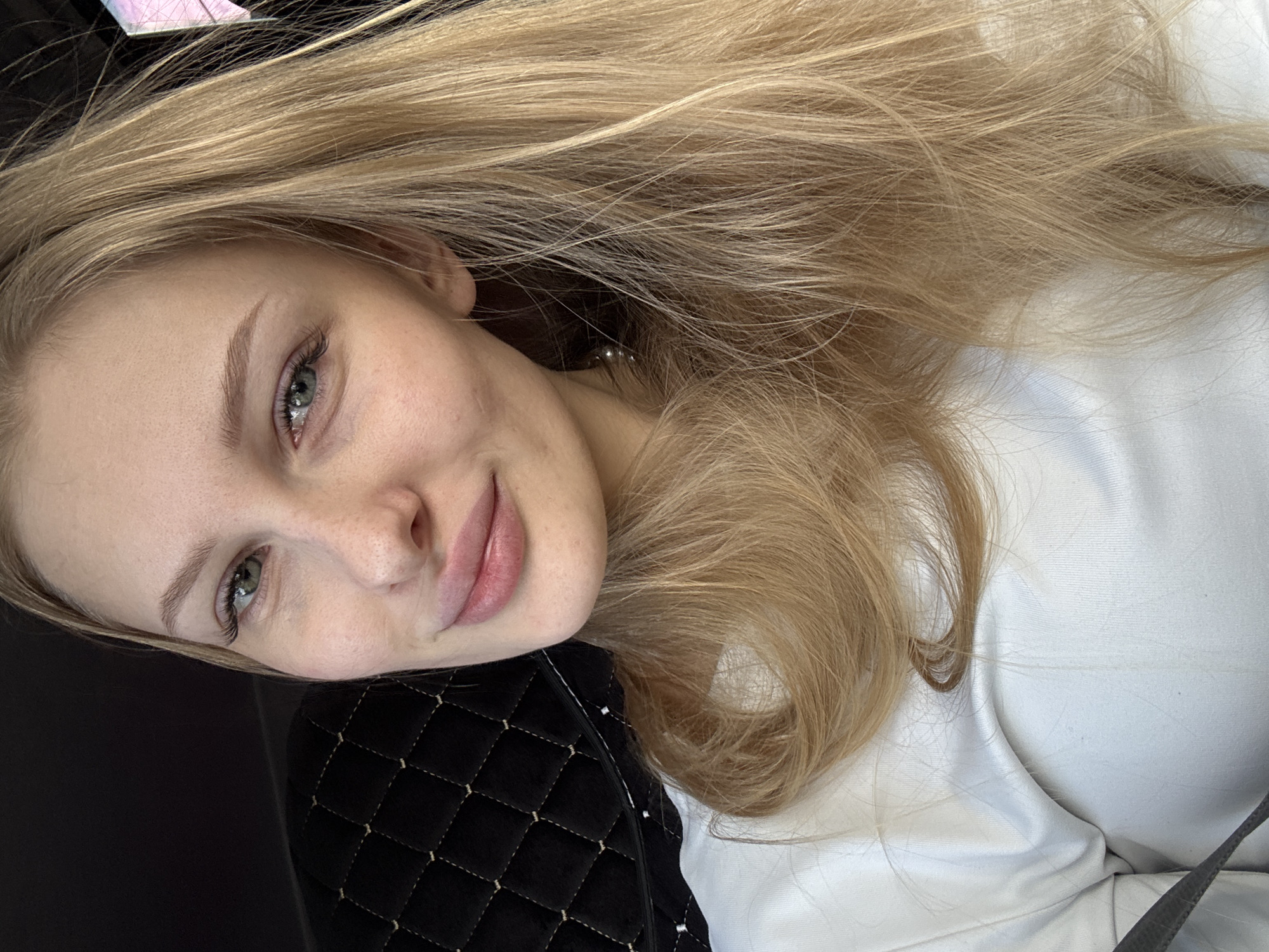 A close-up portrait features a young woman with long, flowing blonde hair, spread out around her head. She is positioned lying down, with her head tilted to the left, resting on a dark, quilted surface. Her gaze is directed slightly upwards and towards the viewer, revealing light-colored eyes, possibly green or blue-green. Her face has fair skin, natural eyebrows, and light pink lips curved into a subtle, gentle smile. Faint skin texture is visible, suggesting minimal makeup.
She is wearing a white top or blouse, visible on her right side. The dark quilted surface her head rests on could be a cushion, pillow, or part of a piece of furniture. A subtle, small earring appears near her left earlobe. The background consists of dark areas, likely behind her head, and brighter white fabric or surface to her right. A hint of a pink object is barely visible in the upper left corner of the frame. The overall scene suggests a relaxed, indoor setting, possibly in Perm, Russia, with soft, ambient lighting. There is no visible text in the image.