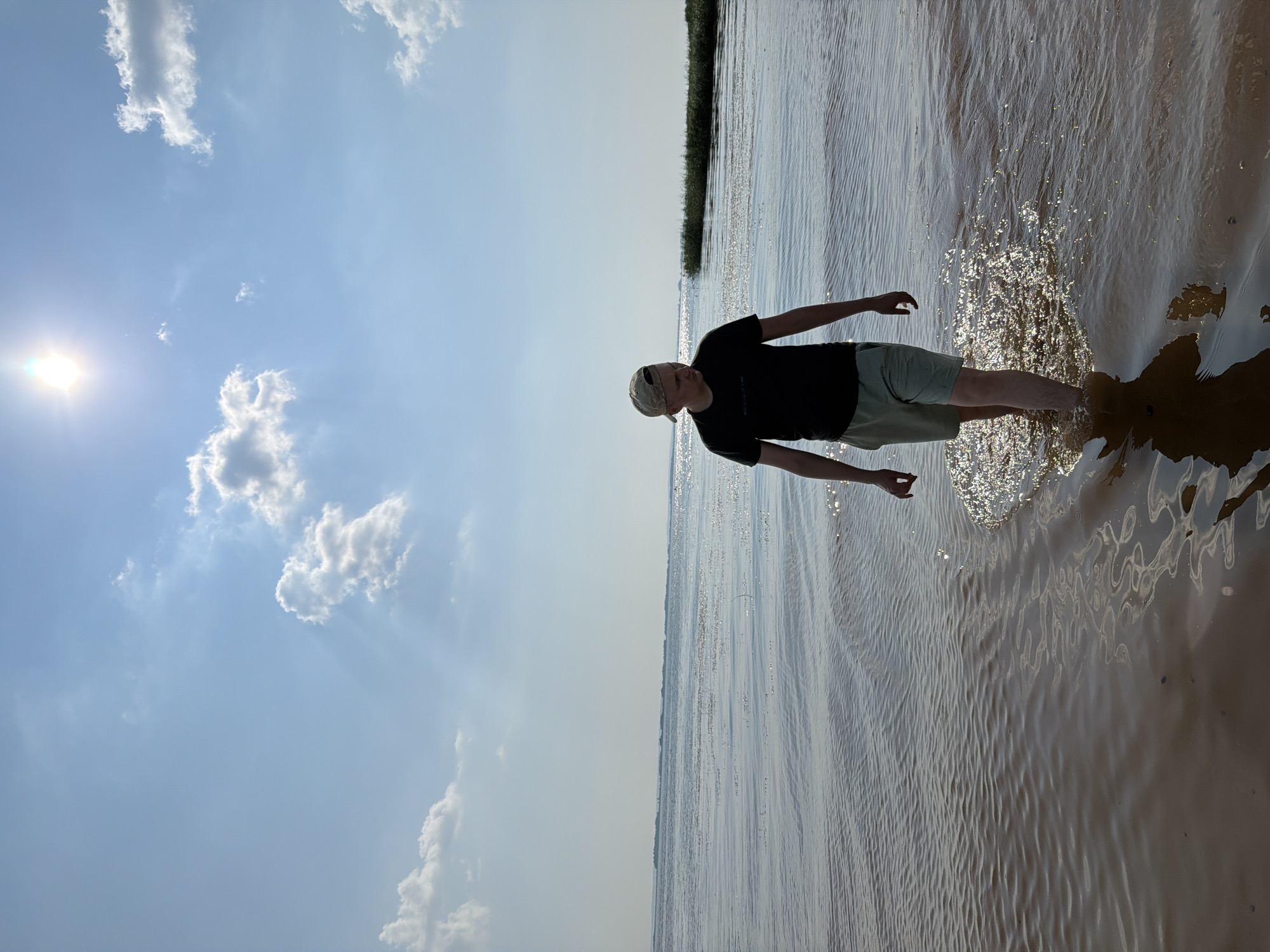 A young man wearing a black t-shirt, light-colored shorts, and a camouflage baseball cap stands in shallow, rippling water. The sun is bright and low in the sky, casting a lens flare on the left side of the image and reflecting off the water's surface, creating a shimmering effect. The water is muddy brown, and the man's legs are partially submerged. His left hand is held up, fingers splayed, near the water's surface, as if he is interacting with it. His right arm is bent at the elbow, with his hand resting on his hip. The background shows a hazy horizon line with what appears to be tall grass or reeds. The sky is a pale blue with scattered white clouds. The image is oriented vertically. The overall scene suggests a warm, sunny day, possibly late afternoon given the sun's low position. The man's posture and the playful interaction with the water hint at a relaxed and enjoyable moment. The location is identified as Krasnyi Bor, Russia.