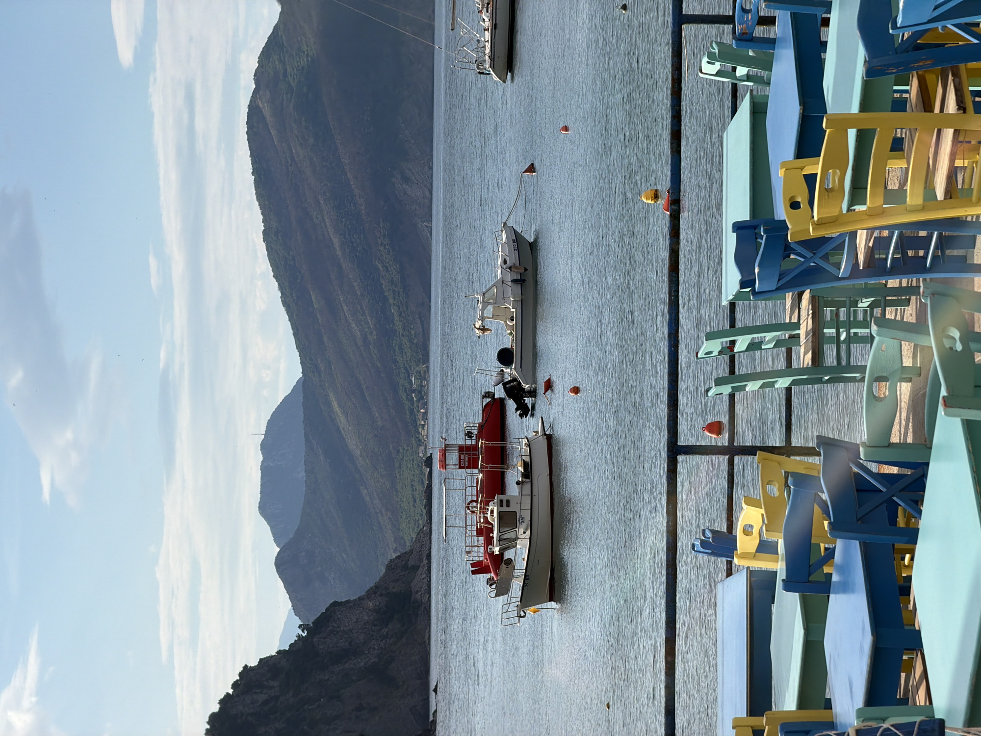 The scene depicts an outdoor setting near the coast, possibly in Petrovac centar, Montenegro. Mountains are visible in the background beneath a sky with scattered clouds. A body of water contains several boats, one of which is red and white. In the foreground, there are tables and chairs of various colors including yellow, blue, and light green, suggesting an outdoor dining area.