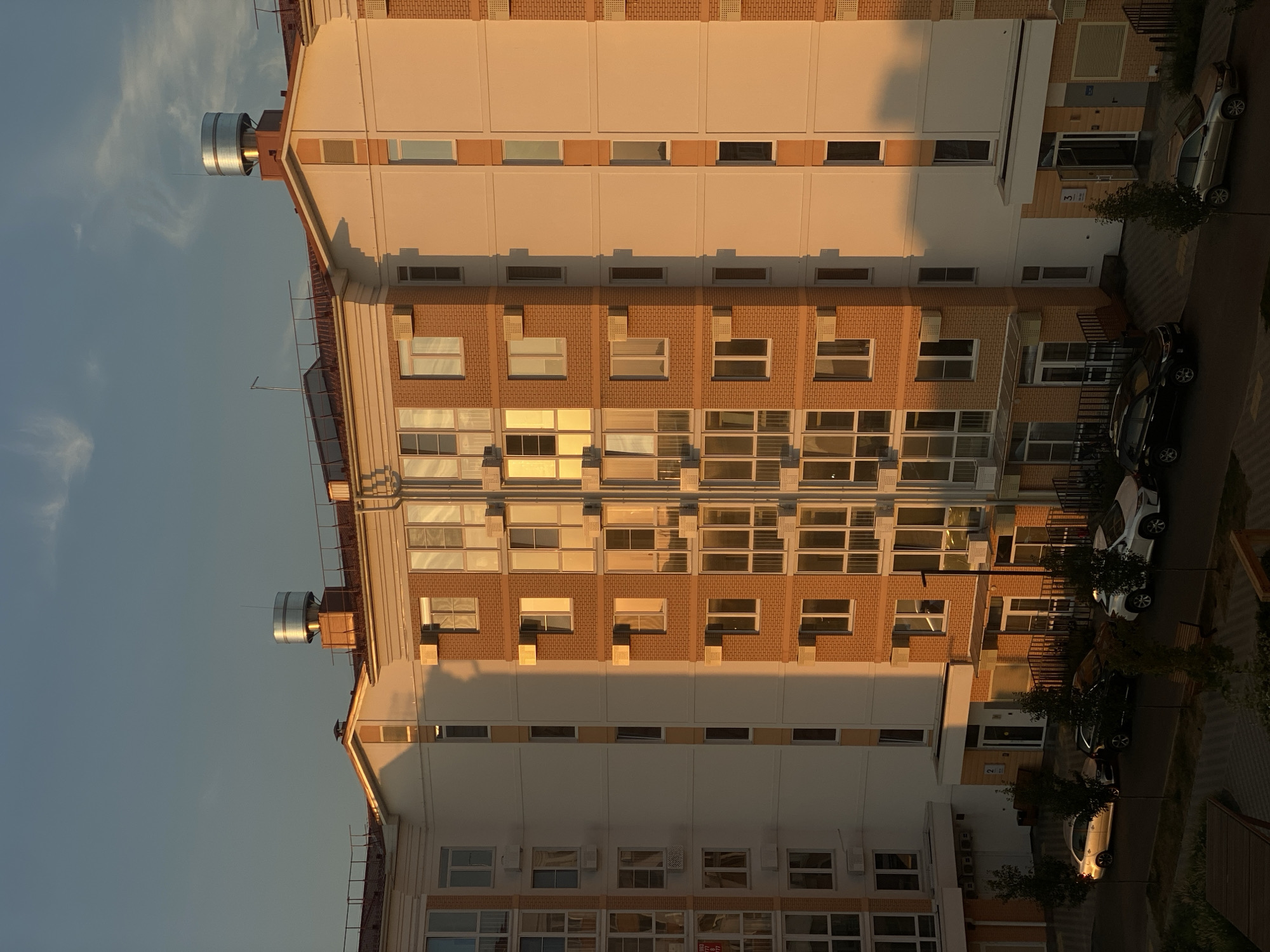 The photograph captures an apartment building in Vishnyaki, Russia, illuminated by the warm glow of sunset. The building features a facade of light-colored plaster and brickwork, with numerous windows reflecting the sky and golden light. Several air conditioning units are visible mounted on the exterior walls. The building has a multi-pitched roof with a large cylindrical metallic structure atop one section. In the foreground, a street or courtyard area is visible with several parked cars, including a silver sedan, a dark SUV, and a white SUV. Trees and benches are also present in the outdoor space. The sky above is a gradient of blue and pale yellow, with wisps of clouds.