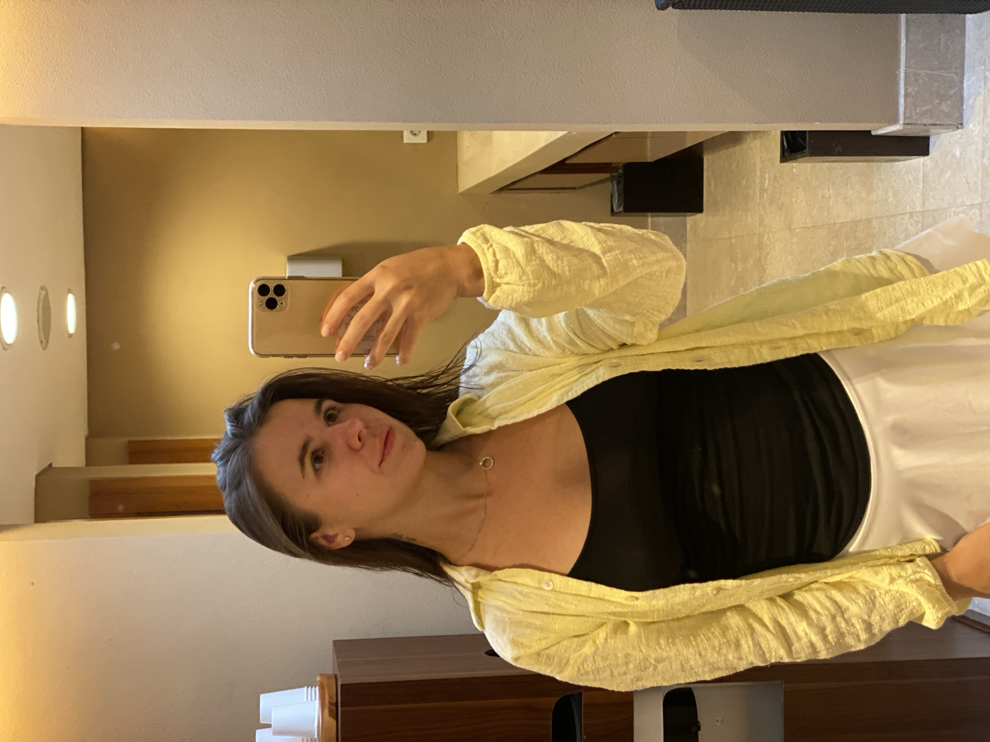 A young woman stands in a bathroom, taking a mirror selfie. She is wearing a black tank top, a yellow open button-up shirt, and a white skirt. She has dark brown hair and is wearing a necklace. The bathroom has beige walls, a tiled floor, and a vanity with a sink in the background. A trash can is visible in the corner of the room. The woman is holding a smartphone to take the picture. The image appears to be taken in Kuta Selatan, Indonesia.
