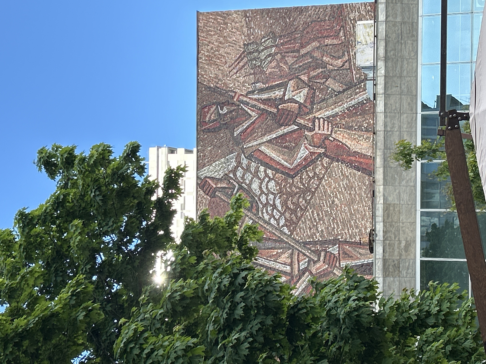 The image shows a mosaic mural on the side of a building, partially obscured by a large green tree in the foreground. The mural depicts figures engaged in what appears to be agricultural or industrial work, possibly related to communism, judging by the style and imagery. The building is modern, with glass windows and concrete or stone cladding. The sky is blue. The location appears to be urban, as indicated by the architectural style.