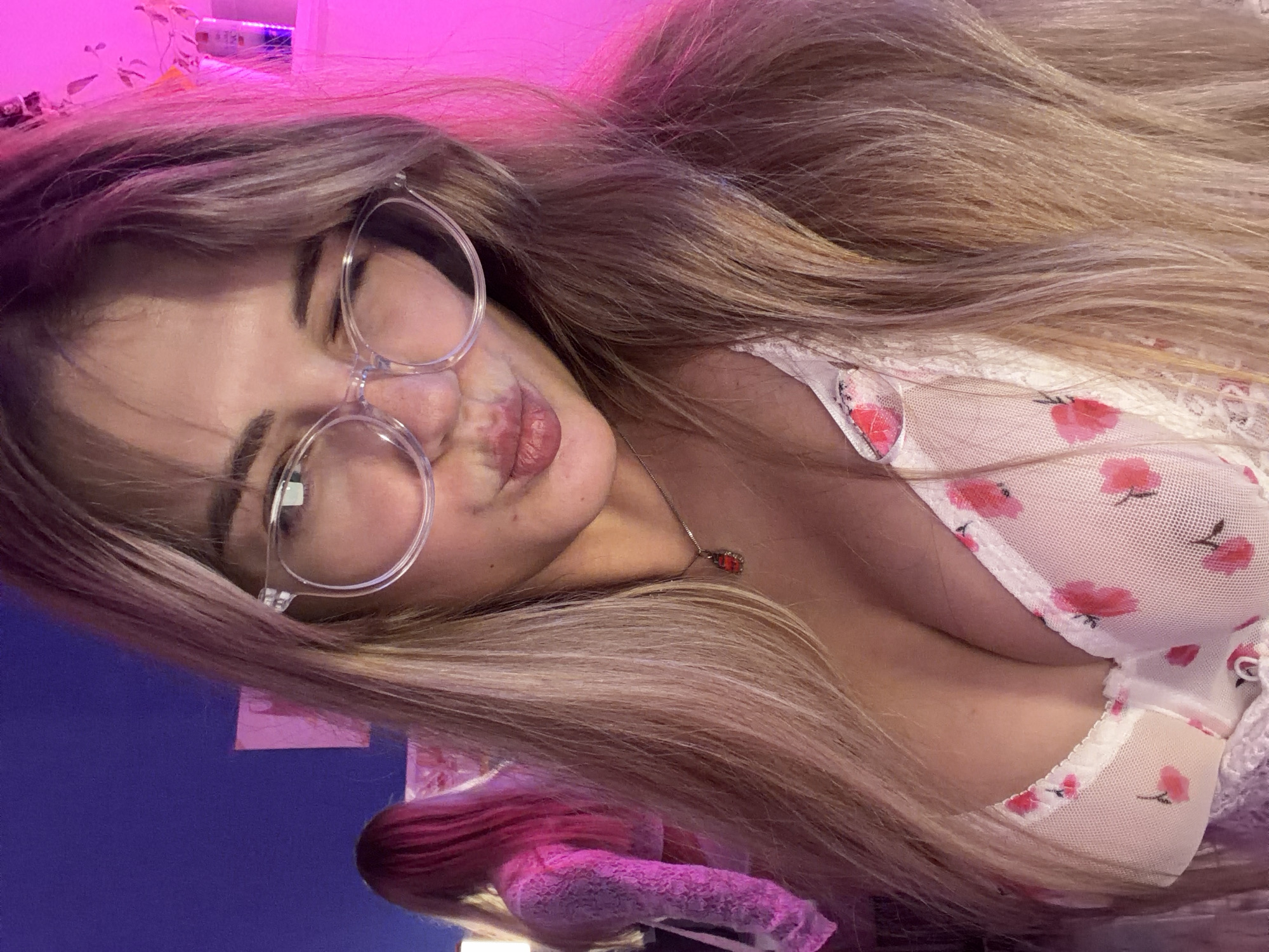 The image is a selfie of a young woman with long blonde hair and wearing clear-framed glasses. She has on a white top with a floral pattern and is pursing her lips. She is wearing a necklace with a red pendant. The background is a mix of purple and pink lighting. It is not possible to ascertain the location in the city of Большая Шильна, Russia from the image.