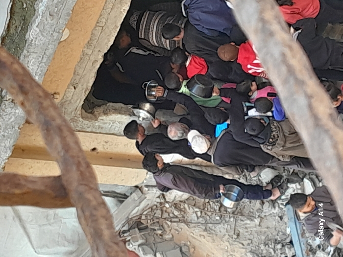 The image portrays a crowded scene in Khan Yunis, Palestinian Territory, set within a severely damaged urban environment. Visible are approximately 15-20 individuals, a mix of adults and children, densely packed around a dark, recessed opening in a structure, possibly a damaged building entrance or basement access point. The surrounding ground and background consist of extensive concrete rubble, debris, and exposed structural elements. Many individuals are actively engaged in passing or receiving metallic pots and bowls, suggesting a distribution or collection of provisions. A man on the left stands on rubble, holding an empty silver pot. Children are interspersed throughout the crowd, some also holding small containers. The scene depicts an organized, urgent communal interaction for resource acquisition. A "Galaxy A14" watermark is present in the lower-left corner.