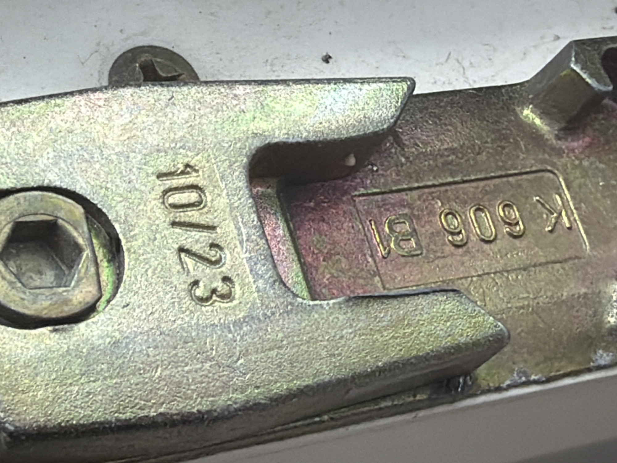 This close-up, eye-level media file shows a metallic, possibly brass or gold-colored object. The object appears to be a component of a mechanical device, such as a lock or latch mechanism, given its shape and the presence of a hexagonal bolt head. The surface of the object is textured, showing signs of wear and age, with some discoloration and a slightly rough finish.
There are two distinct sets of embossed text visible on the object. On the left side, oriented vertically, is the text "10/23". To the right of this, and oriented horizontally within a recessed rectangular area, is the text "K 606 B1". The lighting suggests it is indoors, possibly in a workshop or a room with artificial light, as there are no indications of natural light sources or shadows that would suggest a specific time of day or weather conditions. The overall impression is one of detail and utility, focusing on the markings and construction of a functional part. No people or animals are visible.