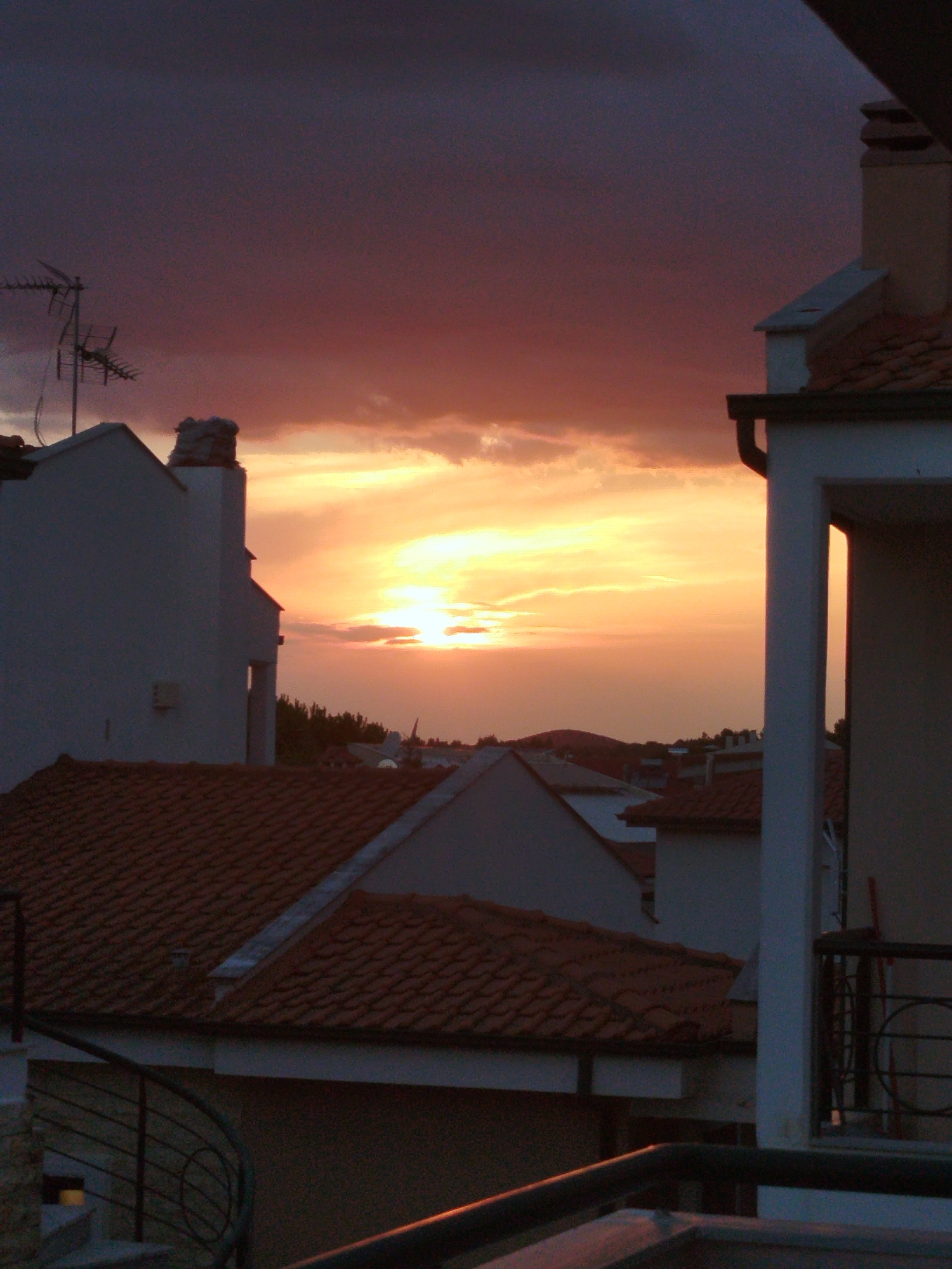 The image depicts a sunset over a coastal town in Sithonia Municipal Unit, Greece. The sky is filled with dramatic clouds tinged with shades of orange, yellow, and deep purple. In the foreground, several rooftops with red tiles are visible, characteristic of Mediterranean architecture. Buildings with white walls and angular designs are interspersed among the roofs. An antenna is prominent on the left side of the image, suggesting a residential area. The overall impression is one of a peaceful, dusk scene in a picturesque Greek locale.