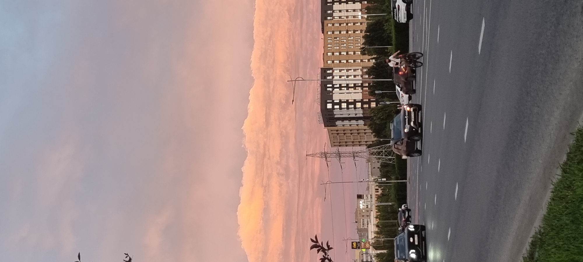 The image depicts a city street scene in Aktobe, Kazakhstan, during what appears to be sunset. The sky is filled with soft pink and orange clouds, indicative of a sunset. In the foreground, a multi-lane asphalt road is visible with white lane markings. Several cars are driving on the road, including a black SUV on the left and a dark colored SUV in the center lane. To the right of the dark SUV, a person is riding a bicycle. In the middle ground, there are trees and a prominent electrical pylon. Behind the pylon, a modern, multi-story building with numerous windows is visible. The overall impression is a typical urban environment at dusk, with traffic and residential or commercial structures under a colorful sky.