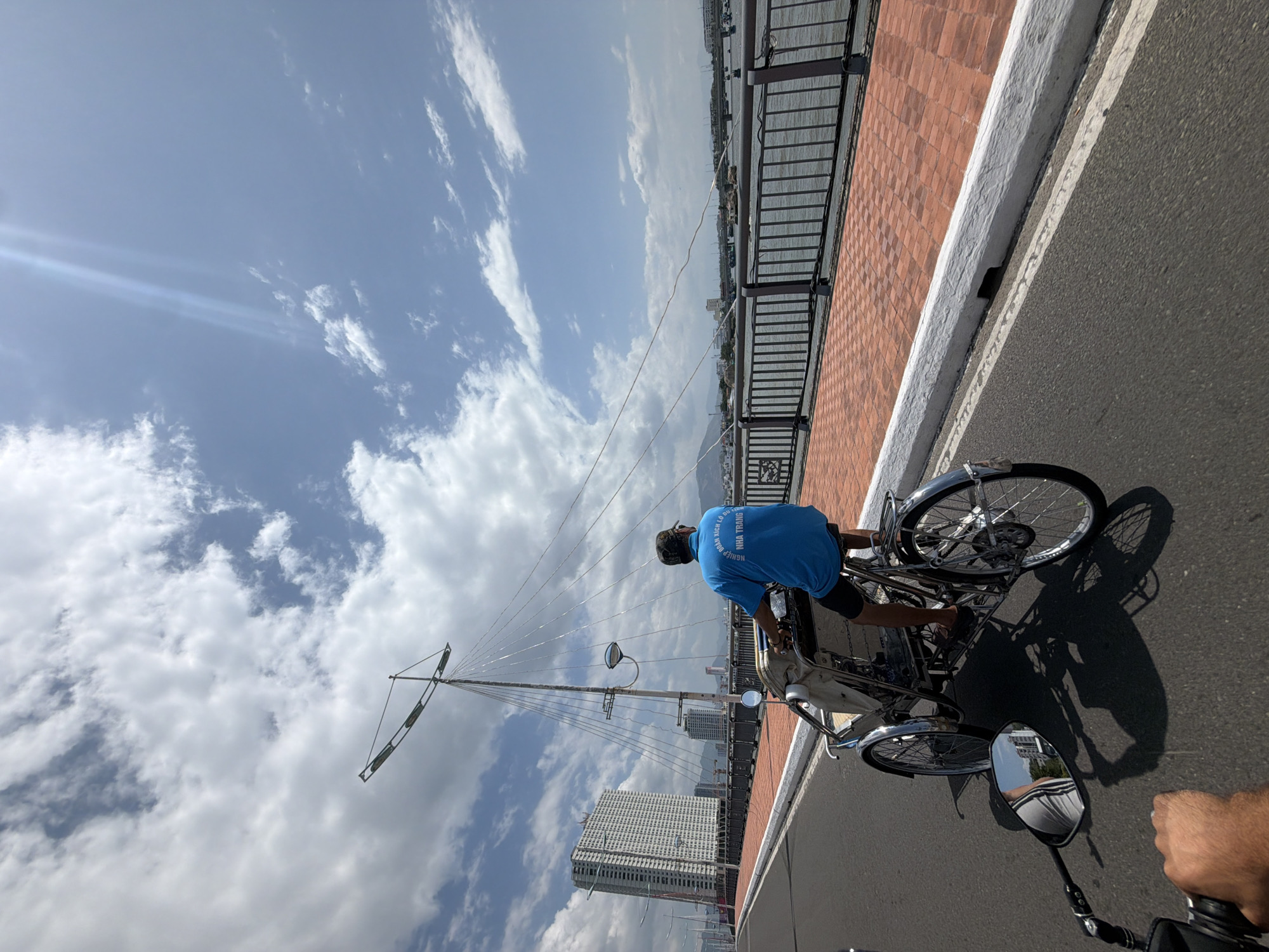 This image, captured from a low angle on a bright, sunny day in Bac Nha Trang Ward, Vietnam, depicts a moment of travel in an urban coastal setting. The primary subject is a person, viewed from behind, wearing a vibrant blue short-sleeved shirt with Vietnamese text printed on the back. The visible text includes "NGHIỆP ĐOÀN" (meaning "Trade Union" or "Syndicate") and "NHA TRANG," indicating a local affiliation with the city of Nha Trang. This individual is operating a tricycle-like vehicle, possibly a pedicab or a motorized rickshaw, featuring chrome accents and fenders.
The photograph appears to be taken from another vehicle, suggested by a rearview mirror and a visible hand in the foreground, indicating the photographer is likely on a motorbike. Both vehicles are traveling on an asphalt road marked with a white line. Adjacent to the road is a white curb separating it from a reddish-brown tiled walkway. A sturdy black metal railing runs along the edge of this walkway, overlooking a vast body of water, likely Nha Trang Bay or the Cai River.
In the mid-ground and background, several modern high-rise buildings dominate the distant skyline across the water, including one prominent, multi-story rectangular structure. A tall, slender mast-like pole, possibly a decorative street light or part of a port structure, stands out against the sky on the left. The sky is clear blue with scattered white and grey clouds, and a bright lens flare indicates direct sunlight, suggesting it is midday or early afternoon. The strong sunlight casts clear shadows of the pedicab and its operator onto the road. The scene captures daily life and transportation in a bustling Vietnamese city.