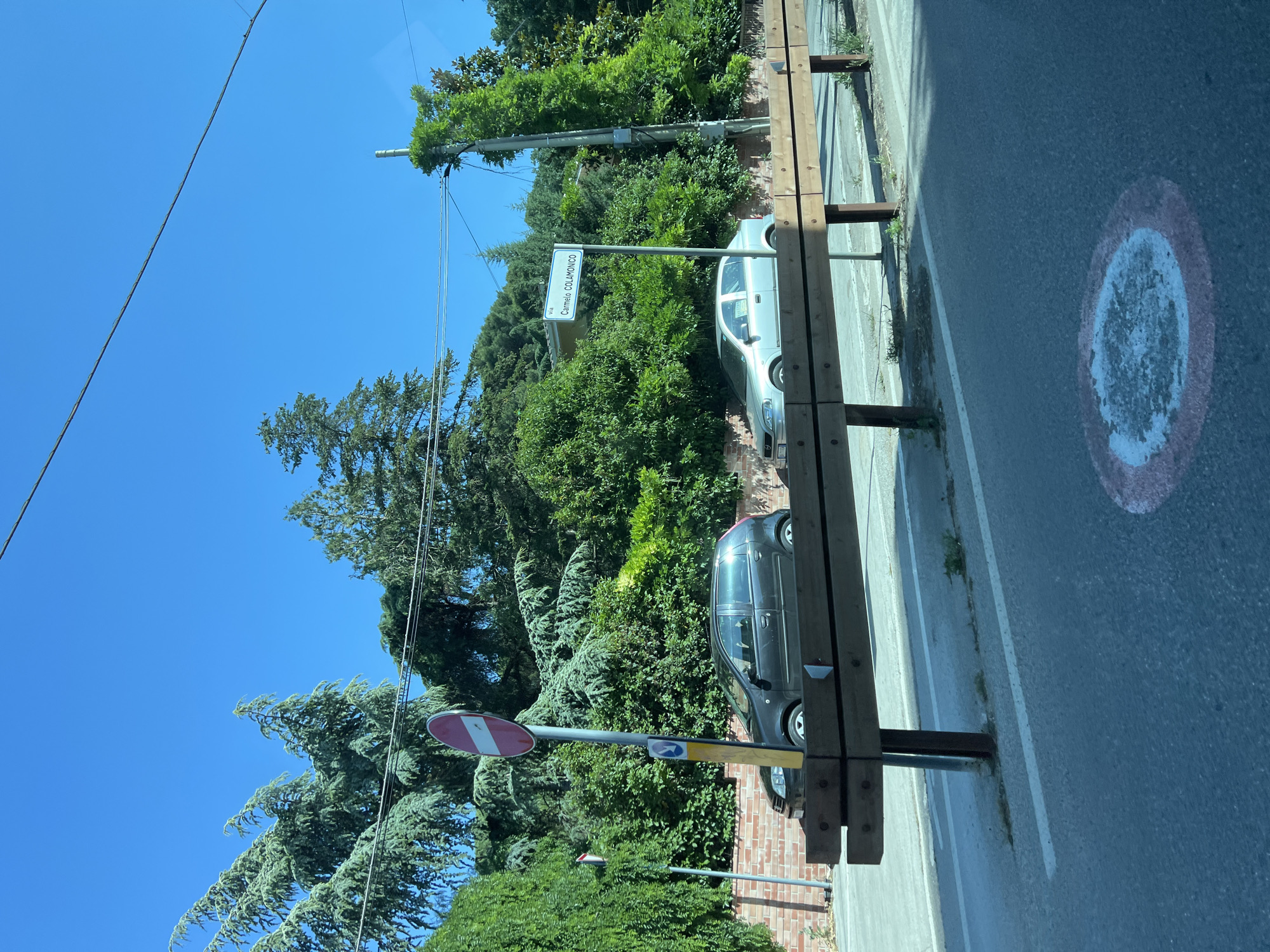 The image depicts an outdoor street scene under a clear blue sky on a bright, sunny day. The location is identified as Via Carmelo Colamonico in Cassano delle Murge, Italy, indicated by a white rectangular street sign with blue text positioned mid-ground on the right.
In the foreground, a dark wooden barrier, constructed from two horizontal planks affixed to vertical metal posts, extends across the lower portion of the image. The road surface is asphalt, featuring faded white dashed lines and a circular road marking with a red outer ring and a white inner area, which is worn.
To the left of the wooden barrier, a tall silver pole supports a circular "no entry" traffic sign (red with a horizontal white bar) and two smaller signs below it, one yellow and one blue. Behind the barrier and signs, a low, reddish-brown brick wall is visible.
Two automobiles are parked on the left side of the road, behind the barrier and against the brick wall. The vehicle closer to the foreground is a dark-colored (appears black or dark grey) hatchback. Further down, a silver-colored hatchback is parked. Both vehicles appear unoccupied.
The background is dominated by dense green foliage, including tall evergreen trees with prominent needle-like leaves and various bushes. A utility pole, partially overgrown with ivy or climbing plants, stands on the far right side of the image, with overhead wires extending across the blue sky. Bright sunlight casts distinct shadows on the road and under the barrier. No individuals are visible in the scene.