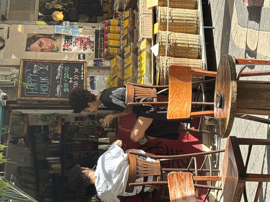 The image depicts an outdoor commercial scene in Tel Aviv-Yafo, Israel, characteristic of a bustling market street such as the Carmel or Levinsky Market. The scene is illuminated by strong daylight, casting distinct shadows.
In the center, two individuals are seated at a bar counter with a vibrant red base. The person on the left, a male with dark curly hair and a white t-shirt, is seen from the back, holding a lit cigarette. He occupies a wooden high stool. The person on the right, a female with dark curly hair in an afro style, wearing a black t-shirt and a dark skirt, is seated in profile, with one leg crossed, and appears to be gesturing with her hands while looking towards the male. She also sits on a wooden high stool. Behind the bar counter, various bottles, glasses, and bartending shakers are visible on shelves.
Above the bar area hangs a blackboard sign with "HAPPY HOUR" in white chalk, accompanied by a martini glass icon. Below, it details "Sunday - Thursday 18:00 - 20:00" and "1+1", followed by Hebrew text that translates to "on all the bar." A security camera is mounted above this sign.
To the right of the bar, an adjacent market stall displays numerous clear plastic bins filled with an assortment of bulk goods, predominantly spices or dried foods, exhibiting yellow, green, and brown hues. Jars with yellow labels are arranged on a higher shelf. Below these displays, several large, woven wicker baskets rest on the ground.
The foreground shows a paved pedestrian area with light-colored pavers and a darker curved section. Two empty wooden chairs, one partially visible, and a small round table made from a repurposed wooden cable spool are positioned. The table holds a small metal ashtray and a metal caddy, possibly for condiments or utensils. The background wall features a large poster of a woman's face with Hebrew text, alongside various other smaller posters and flyers, below an overhead canopy draped with green foliage.