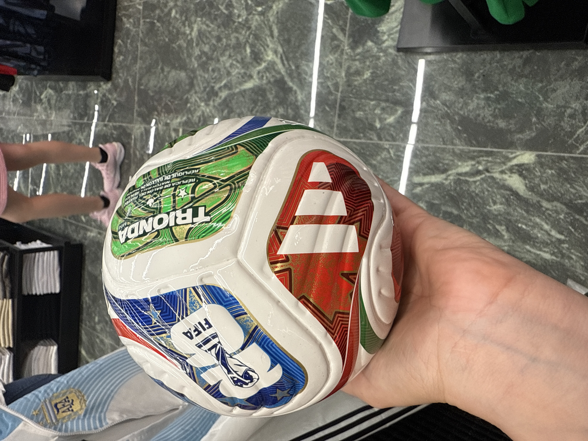 A light-skinned hand holds a new, multicolored soccer ball indoors within a retail establishment in Sunrise, United States of America. The soccer ball, adorned with the FIFA logo and the word "TRIONDA," alongside text indicating "REPLICA MATCH BALL" in multiple languages, features an Adidas logo (three stripes) on one of its red and gold panels. Its design incorporates white, blue, green, and red/gold panels, visually consistent with the Adidas Al Hilm ball from the 2022 FIFA World Cup. The ball's surface appears glossy, potentially from a protective film. In the immediate background to the left, a light blue and white striped fabric with a gold crest, resembling an Argentina national football team jersey, is partially visible. Further back, black display shelves hold neatly folded light-colored merchandise. To the right, the lower legs of a person wearing pink athletic shoes, dark socks, and the bottom edge of a pink garment (skirt or shorts) are visible standing on a dark, glossy marble-patterned floor with white veining and reflections from overhead lighting. A black retail display unit and a partial green object are also present on the right side of the frame.
