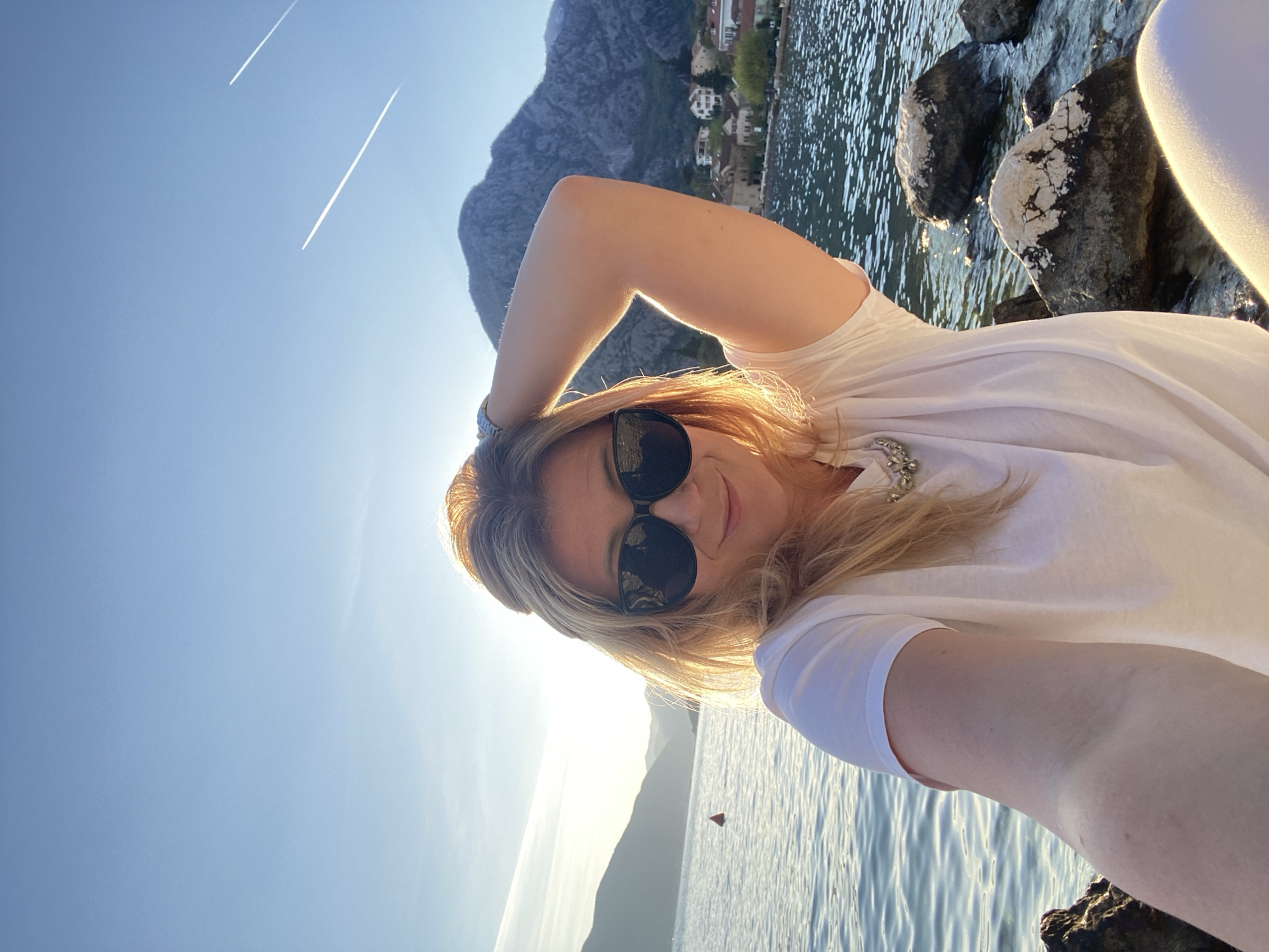 A light-skinned woman wearing sunglasses and a white t-shirt stands near the water in Donji Orahovac, Montenegro. A mountain rises in the background, and a small settlement can be seen along the shore. Contrails are visible in the clear blue sky. The woman has her arm raised, with the sunlight creating a halo effect. Large rocks line the water's edge, and a red buoy is visible in the water.