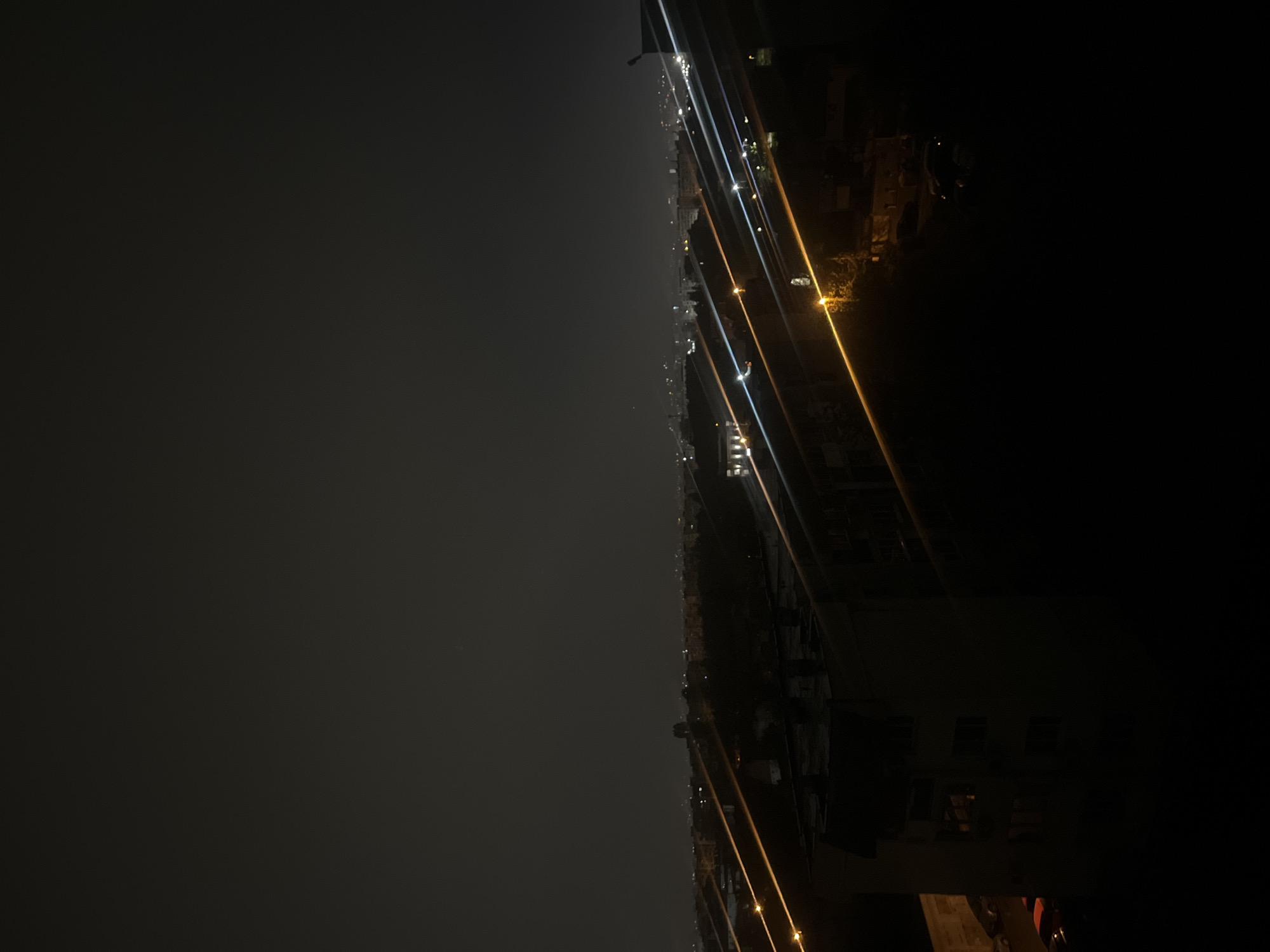 This is a nighttime photograph capturing an expansive urban landscape, likely from an elevated vantage point such as a balcony or rooftop. The image is oriented vertically (originally, the upload is rotated 90 degrees clockwise from how it was captured).
The upper portion of the image is dominated by a dark, overcast sky, appearing as a uniform grey expanse that gradually darkens towards the lower right. A horizontal line, stretching across the middle, marks the distant city skyline, which is dotted with numerous small, diffused lights. These lights, mostly white and yellow, suggest buildings and infrastructure spread across the urban area.
Prominently featured in the lower half and right side of the image are several elongated, diagonal streaks of light. These bright lines, varying in color from bluish-white to warm yellow and orange, are likely lens flares or reflections from nearby artificial light sources, such as streetlights or building illuminations. They add a dynamic element to the otherwise still nocturnal scene.
In the bottom right corner, the dark, silhouetted forms of foreground structures are visible. These indistinct shapes, possibly parts of a building or railing close to the camera, are largely obscured by shadow, with only minimal light revealing faint details.
The time of day is clearly night, and the sky's appearance suggests overcast or hazy weather conditions, which diffuse the distant lights. No specific activities or individuals are discernible, nor is any text visible. The setting is an urban environment, consistent with a city like Sofia, Bulgaria.