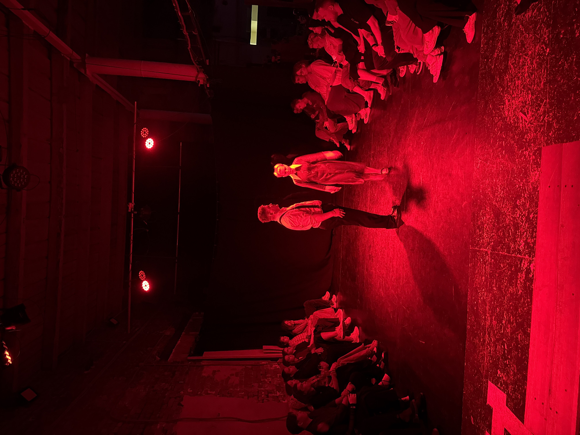 The image shows a theatrical performance illuminated by red stage lighting. Two performers are centered on the stage. A man in trousers and a shirt stands next to a woman in a dress. Seated audience members surround the stage on two sides. The location is possibly a theater in Izhevsk, Russia, but this detail is not discernible from the image itself.