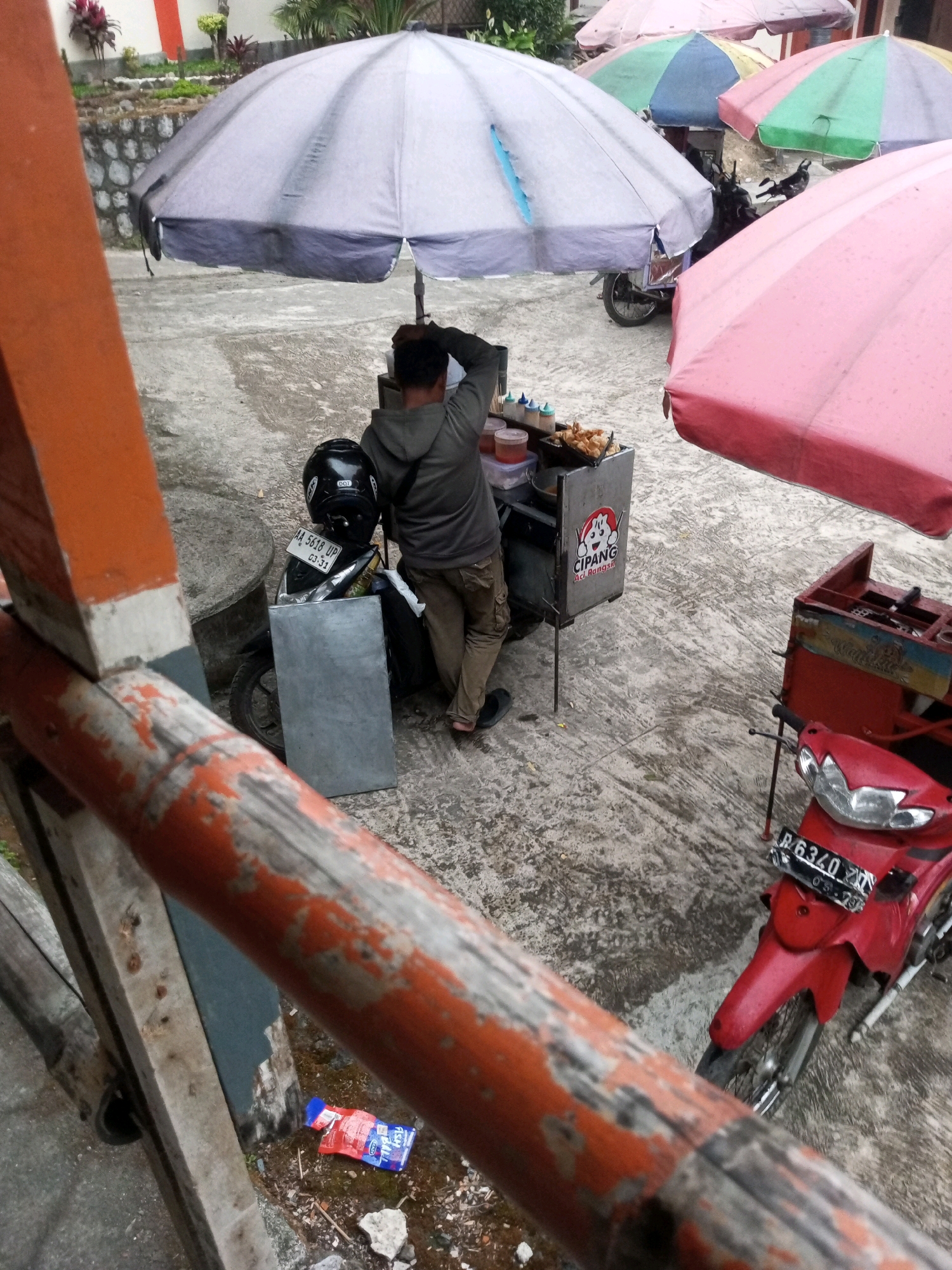 A vendor is preparing food under a large umbrella on a paved outdoor area in Kalibeber, Indonesia. The vendor, wearing a gray hoodie and khaki pants, is facing away from the camera, with one hand on their head. In front of the vendor is a food cart with a logo that reads "CIPANG Aci Bangkit". The cart contains food items and containers of sauces. Behind the vendor is a motorcycle with a black helmet placed on its seat. Another motorcycle, red in color, is parked to the right of the scene. Several colorful umbrellas, pink, green, and blue, are also visible, providing shade. The ground is paved and appears wet in some areas. In the lower left of the image, a red and blue packet is lying on the ground. The environment suggests a street food stall or a temporary market.